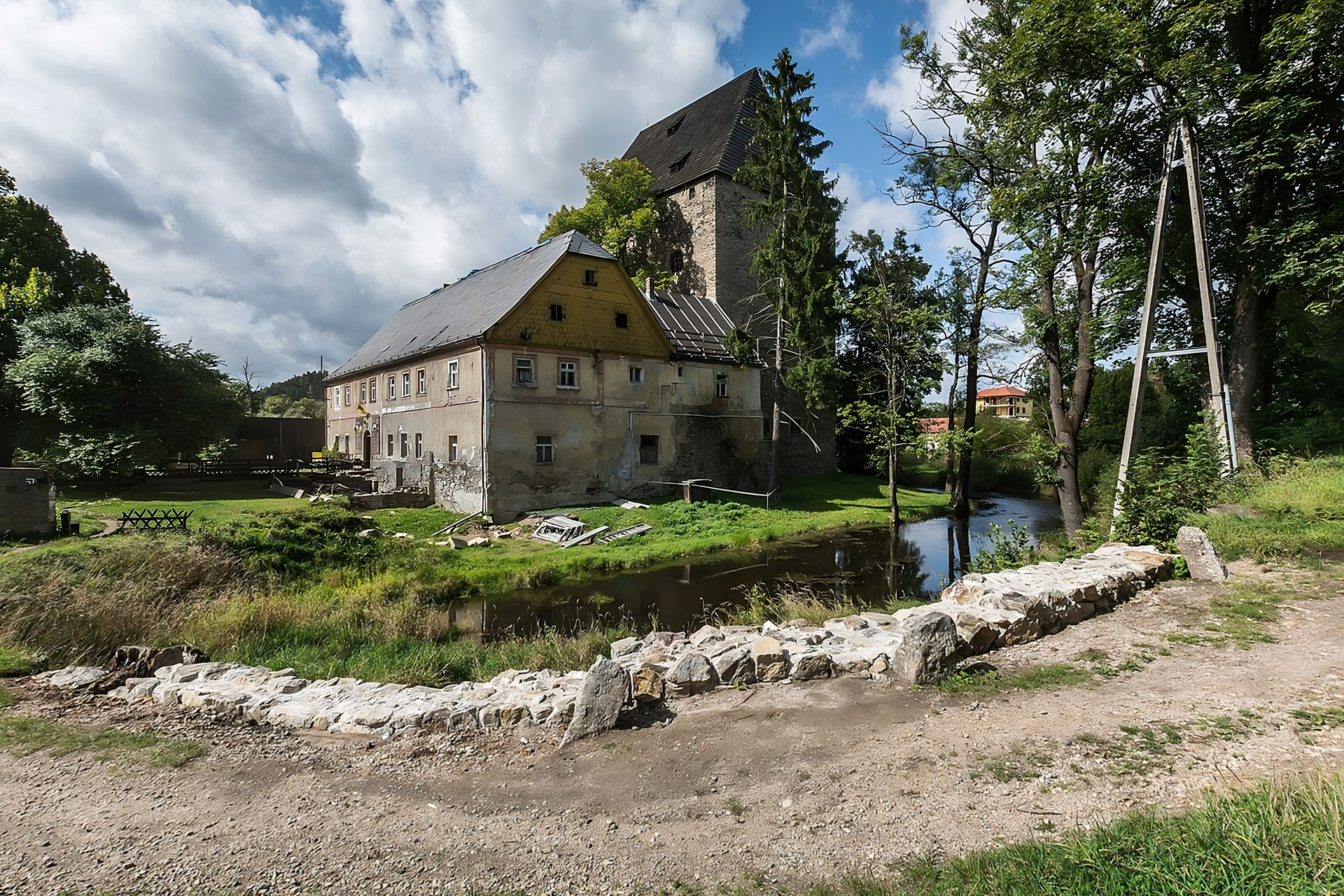 Fotografia przedstawia wieżę w Siedlęcinie. Na pierwszym planie widoczny staw, a dalej porośnięte trawą podwórko. Na podwórku wieża z dobudowanym budynkiem. Budynek, znajdujący się z lewej strony zdjęcia, jest piętrowy i ma skośny dach. Elewacja jest szara. Za budynkiem, z prawej strony zdjęcia, widoczna jest wyższa od budynku, wieża ze skośnym dachem. Wieża wykonana jest z ciemnych cegieł. Wieża częściowo przysłonięta jest zielonymi drzewami iglastymi. Z prawej strony zdjęcia, po drugiej stronie stawu, kolejne drzewa, a także słup z kablem. Niebo jasne, przysłonięte białymi chmurami kłębiastymi.