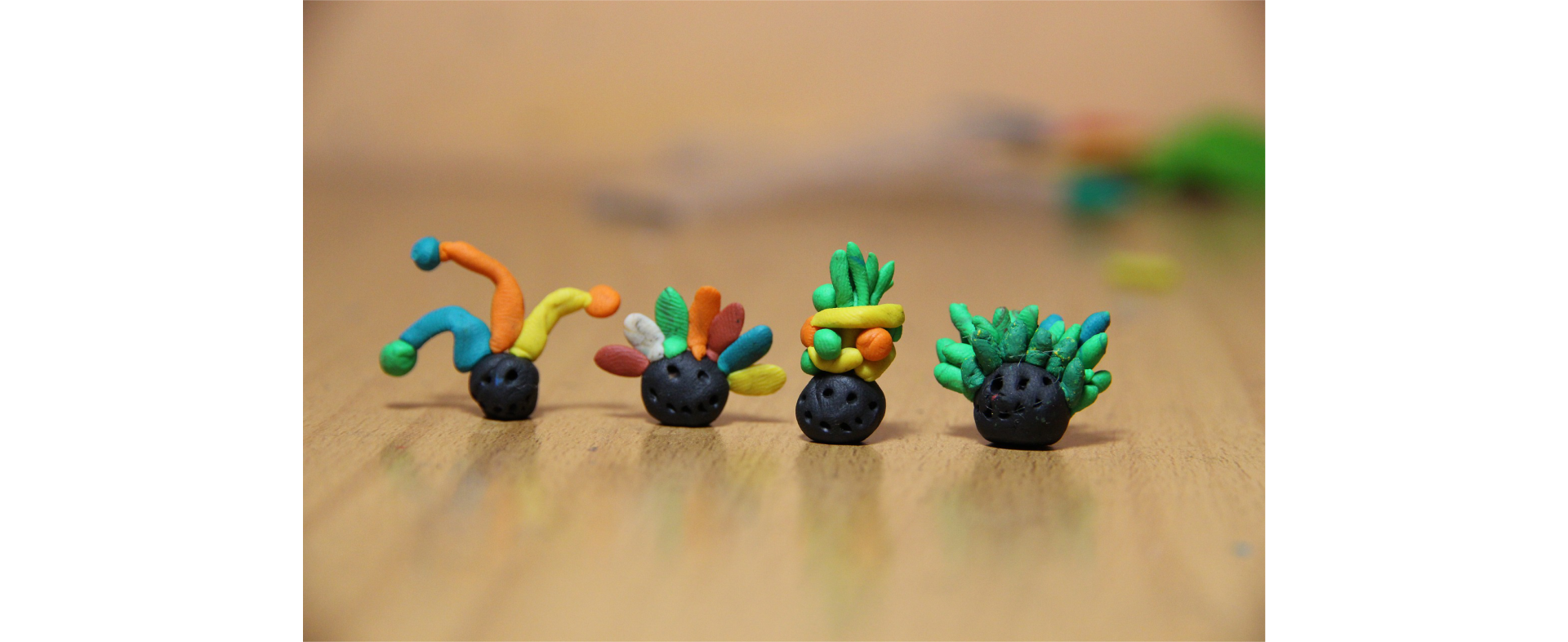 Na stole ustawione są w jednym rzędzie cztery figurki z plasteliny przedstawiające główki z różnymi czapkami. Od lewej: czapka królewskiego klauna, czyli trzy długie części zakończone kulkami, czapka z kolorowym pióropuszem, czapka z owocami, czapka z zielonymi liśćmi.