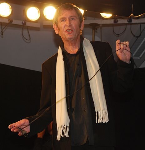 Kolorowa fotografia przedstawia pięćdziesięcioletniego mężczyznę, do pasa, na tle świecących świateł w teatrze. Jest ubrany w czarny garnitur i czarną koszulę, na ramionach ma biały szalik. W ręku trzyma czarny kabel elektryczny. Usta ma otwarte, tak, jakby coś mówił.