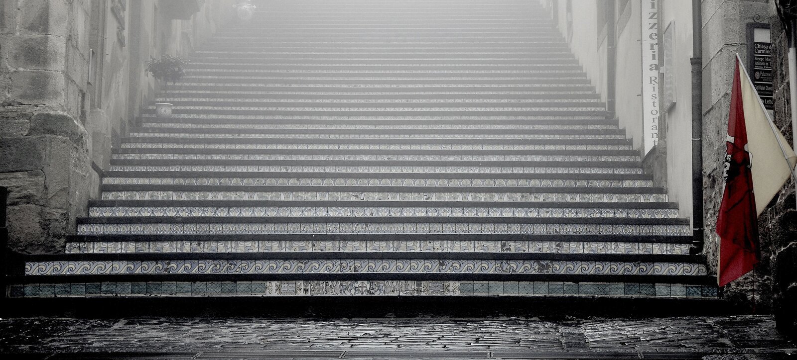 Na zdjęciu widoczne są szerokie schody. Każdy stopień zdobi wzór. Wzory są na kafelkach, którymi wyłożone są schody. Zdjęcie zostało zrobione u ich dołu. Po prawej i lewej strony schodów stoją ściany budynków.