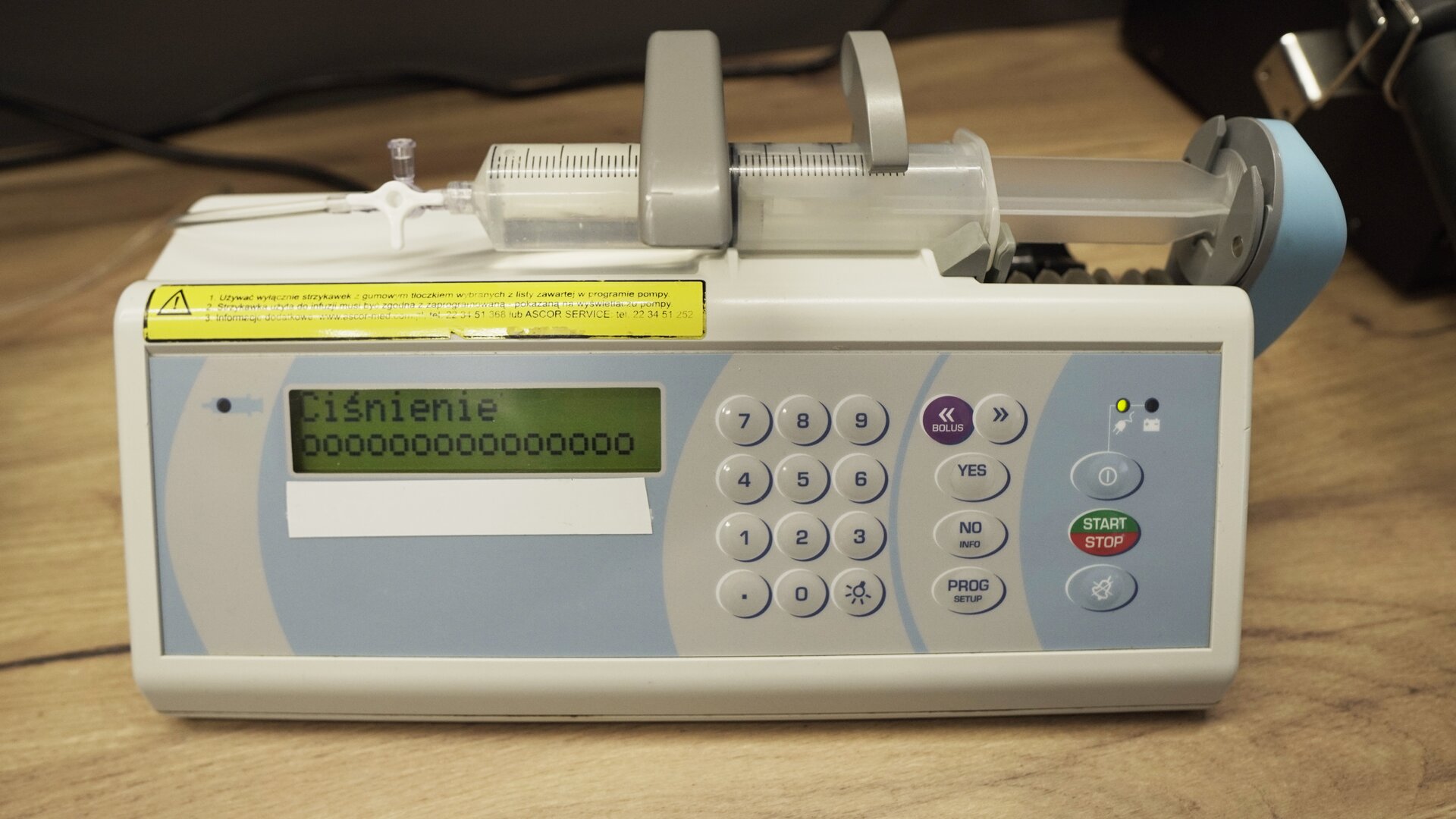 Na zdjęciu strzykawka wypełniona wodą znajduje się w ramieniu pompy infuzyjnej. Na ekranie aparatu wyświetlana jest informacja: "Ciśnienie", a pod spodem 15 okręgów. 