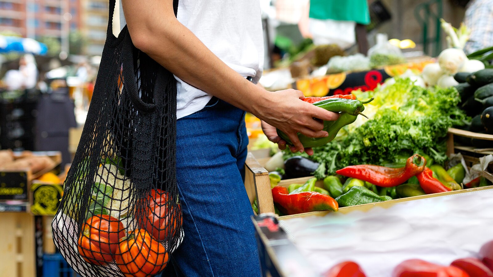 Na zdjęciu widoczna jest osoba, która robi zakupy na targu z warzywami. Na ramieniu ma przewieszoną torbę z siatki, w której widoczne są  pomidory i kalafior. W dłoni trzyma paprykę. 