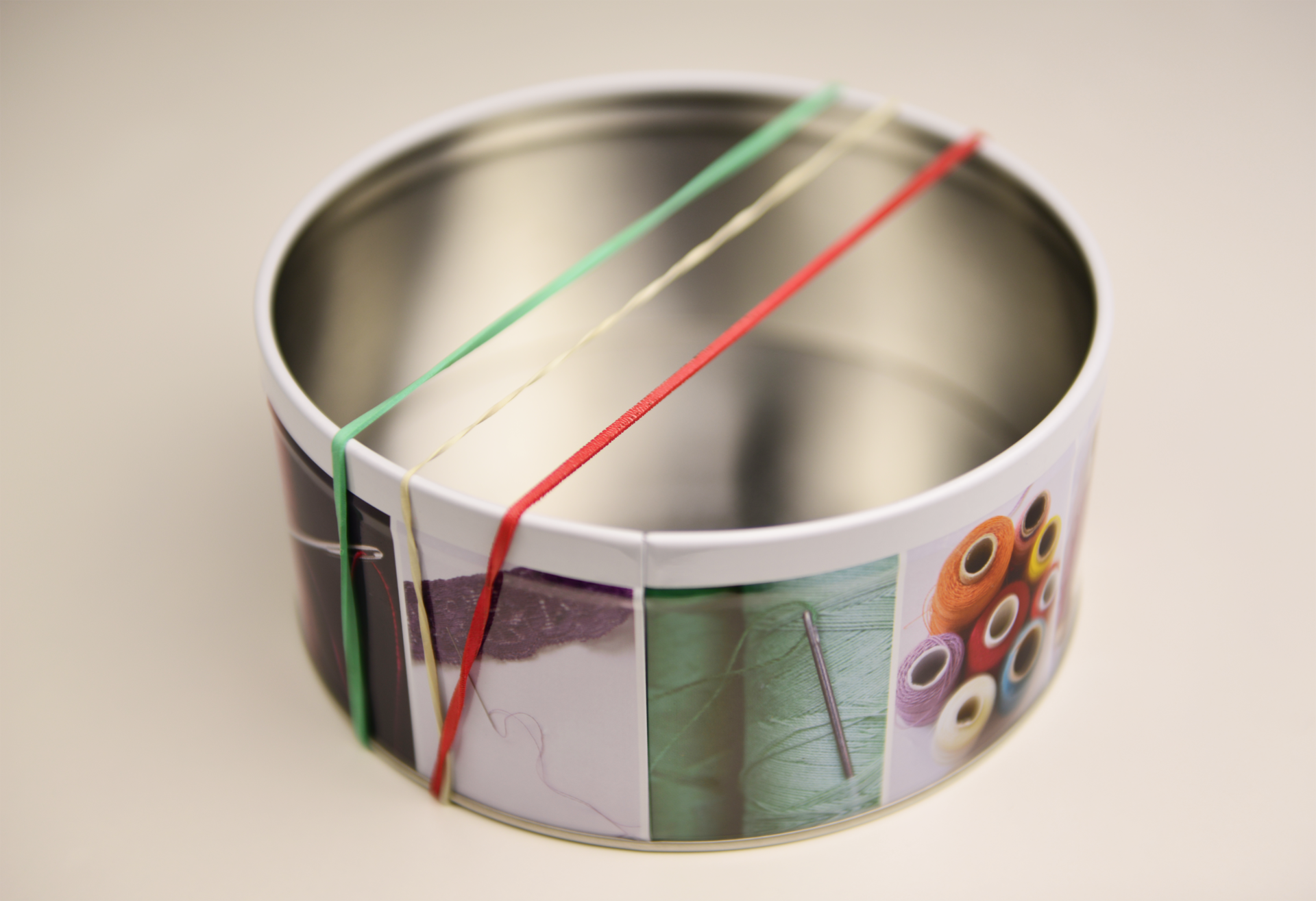 Fotografia przedstawia stół, gdzie postawione jest otwarte, okrągłe metalowe pudełko z różnymi nadrukami. Na pudełko założono trzy gumki: zieloną, białą i czerwoną.