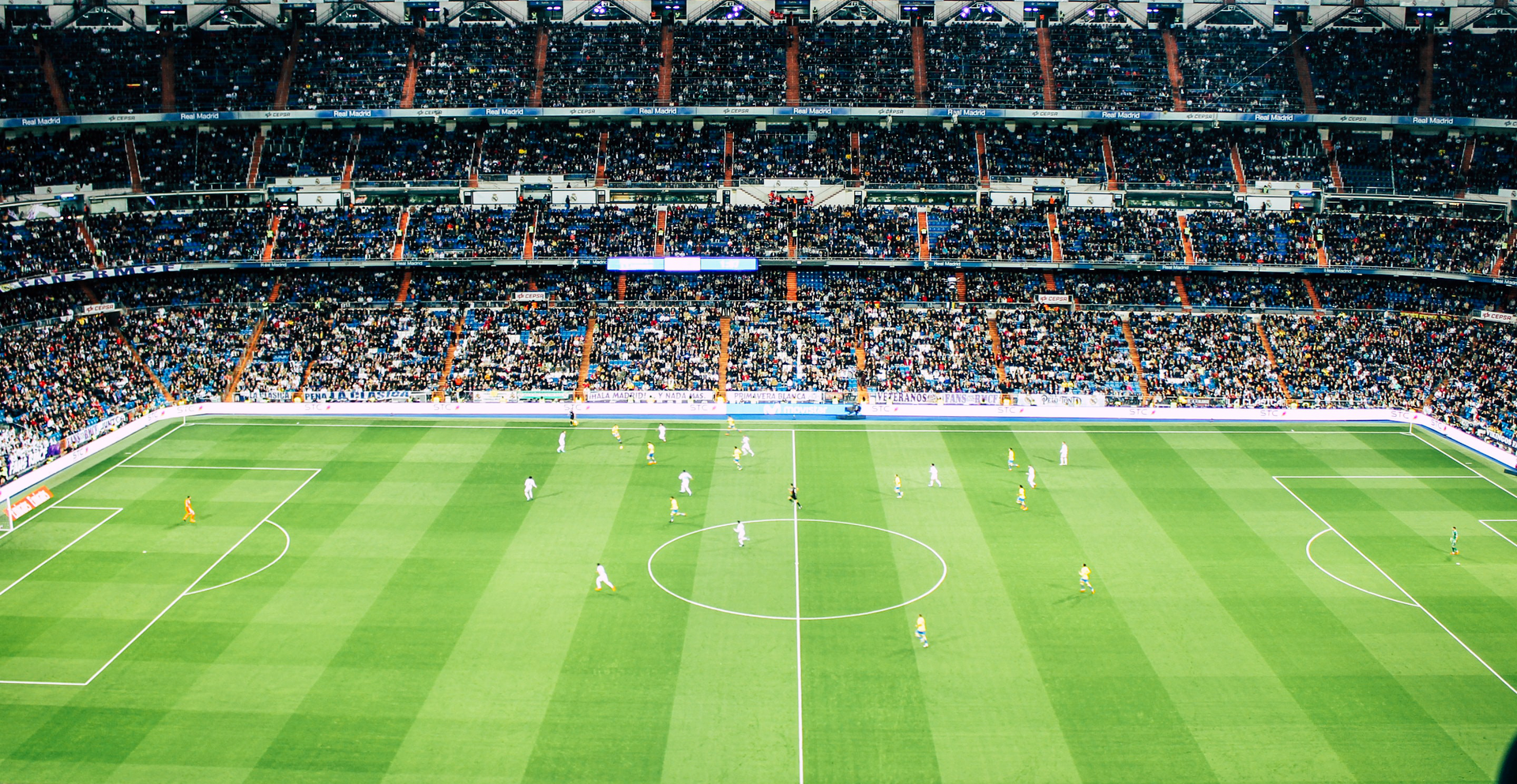 Zdjęcie przedstawia stadion do gry w piłkę nożną pełen kibiców. Na murawie znajdują się piłkarze.
