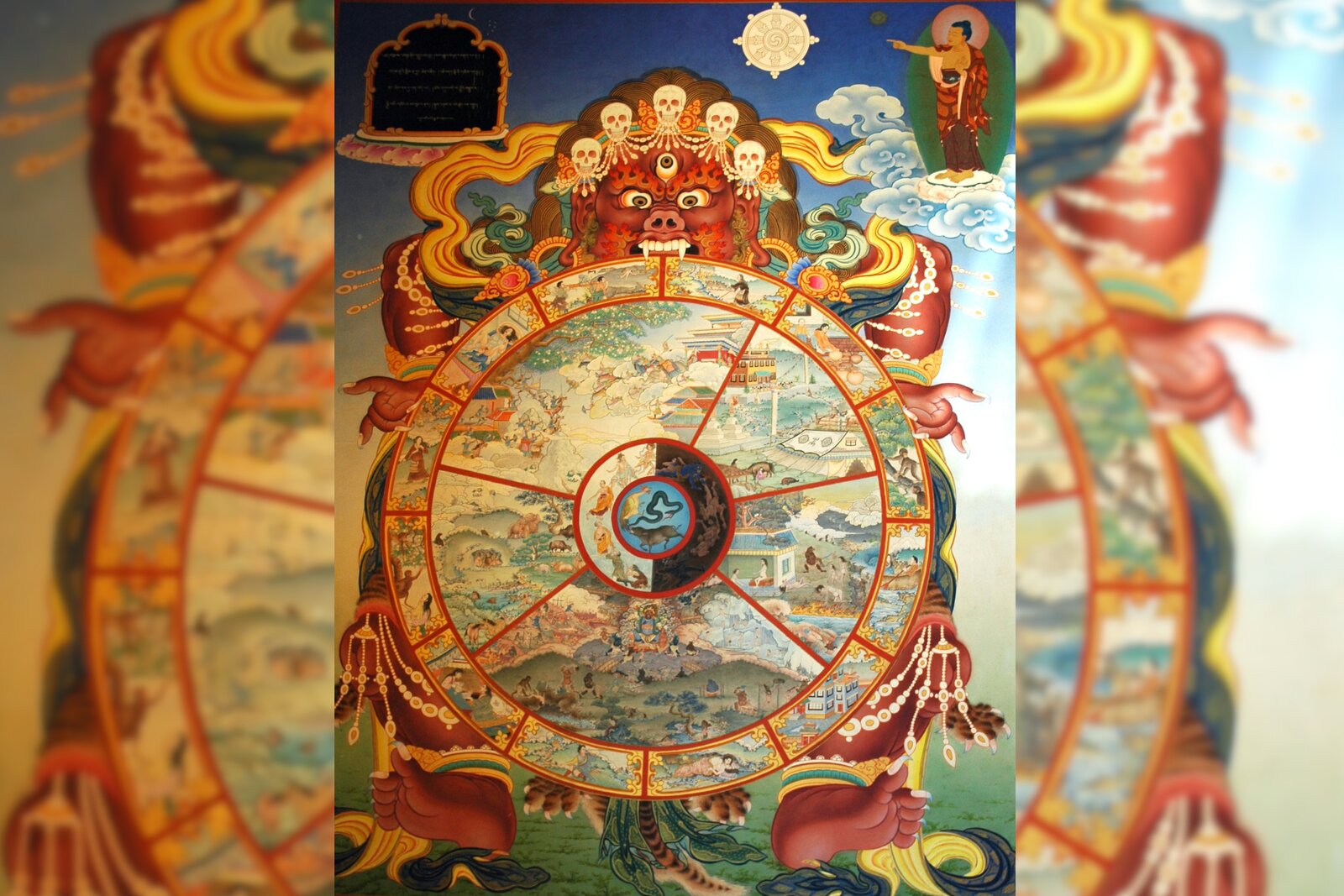 Obraz przedstawia wielkiego czerwonego stwora, który trzyma koło. Na kole znajdują się mniejsze obrazy, które przedstawiają różne sceny. Nad stworem unosi się na chmurze postać wskazująca na symbol buddyzmu.