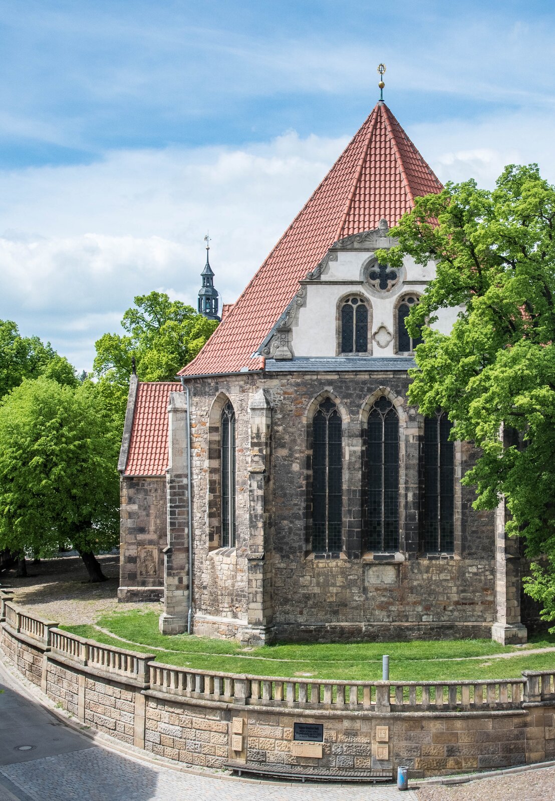 Ilustracja przedstawia tył kościoła w Arnstadt. Kościół jest zwany jako Bachkirche. Duży strzelisty kościół otoczony zielenią.