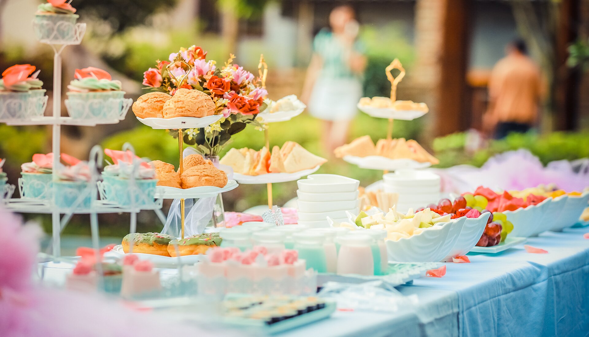 Zdjęcie przedstawia stół nakryty białym obrusem, na którym ustawiono patery z ciastkami, talerze z przekąskami oraz misę z owocami. 