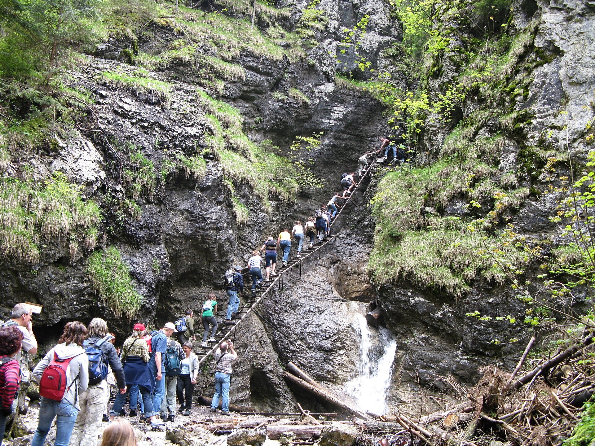 Fotografia turystów wspinających się po stromych schodach umieszczonych na skalnej ścianie, obok której spływa wodospad.