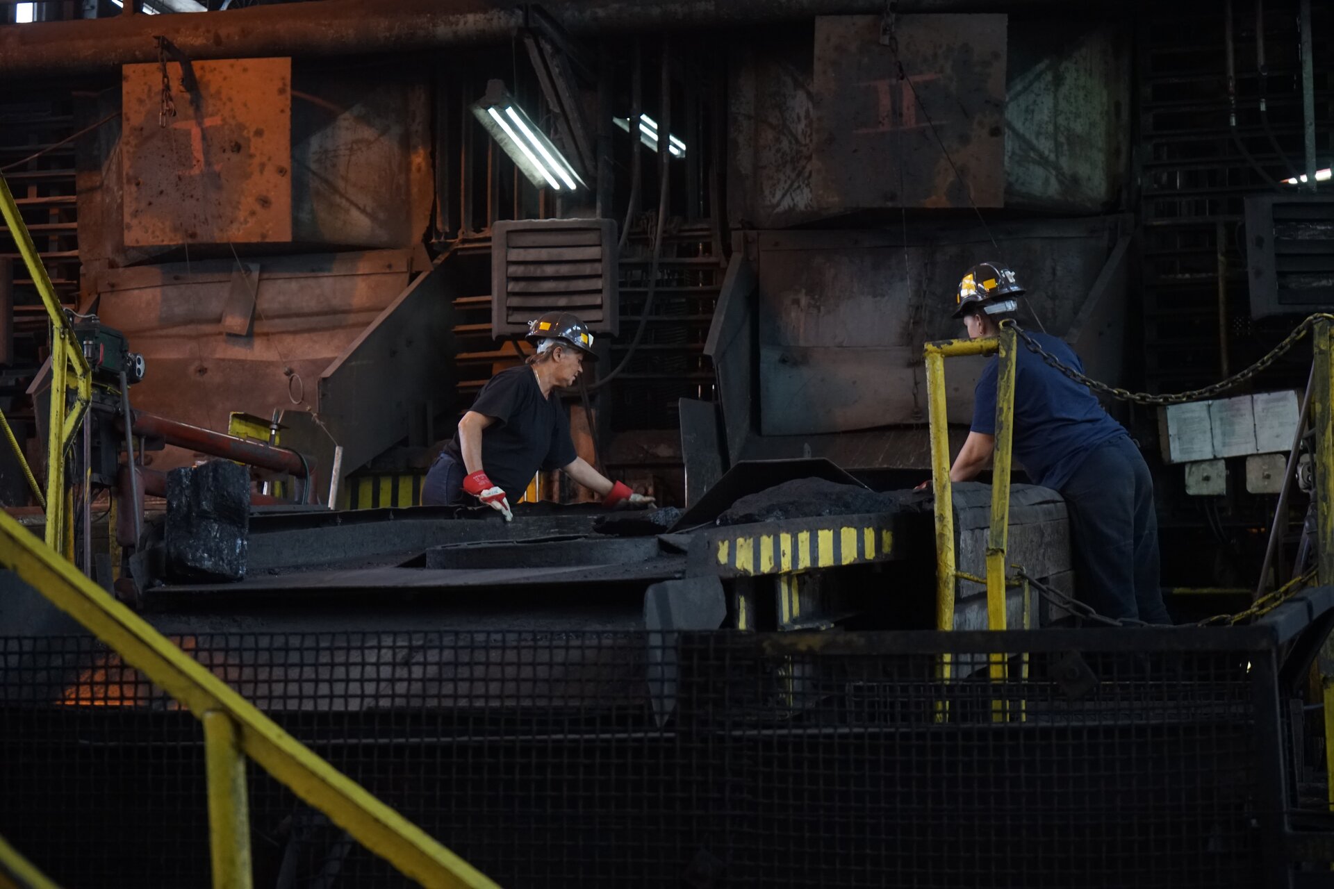 Zdjęcie przedstawia taśmy przebiercze w sortowni zakładu przeróbczego. Trafiają na nie ziarna o większych rozmiarach niż szczeliny sit przesiewacza wibracyjnego. Jego fragment znajduje się po prawej stronie. Ziarna transportowane są od prawej do lewej strony. Po obu stronach przenośnika stoją pracownicy zakładu. Są ubrani w stroje robocze. Mają kaski ochronne, maseczki, rękawice oraz słuchawki wygłuszające. Wybierają materiały pochodzenia niedołowego z taśm. Poniżej zdjęcia widoczny jest przycisk multimedialny z nagraniem audio. Treść nagrania jest zgodna z tytułem zdjęcia.