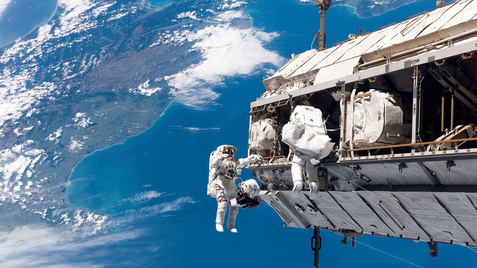 Zdjęcie przedstawia astronautów w skafandrach, obok stacji kosmicznej.