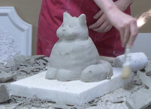 Zdjęcie przedstawia detal architektoniczny wykonany z gliny. Tym detalem jest figurka kota średnich rozmiarów. Pracownik pędzlem czyści powstały odlew.  