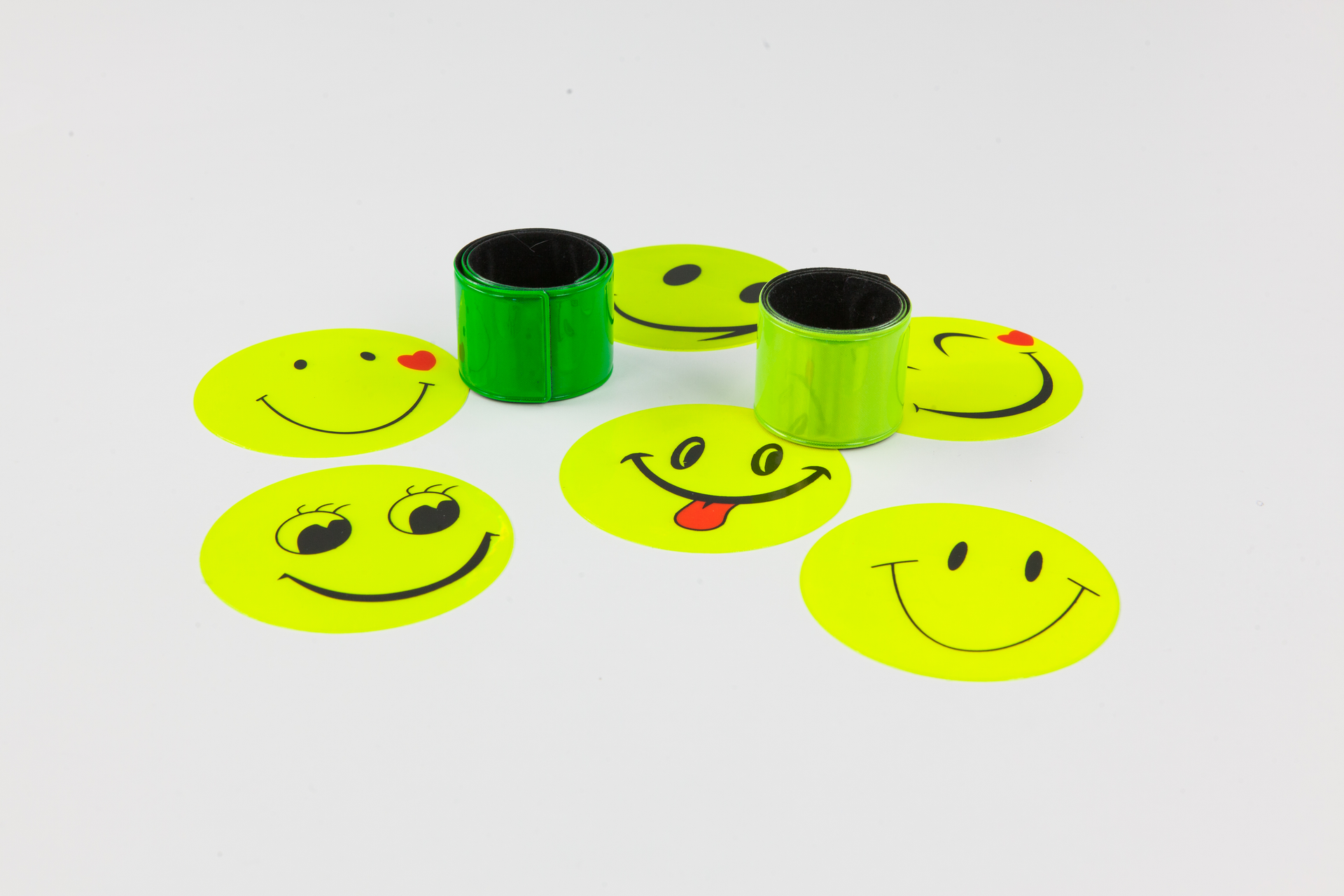 Zdjęcie przedstawia naklejki i opaski odblaskowe. Okrągłe naklejki przedstawiają uśmiechnięte buźki. Elastyczne opaski przeznaczone do zakładania na nadgarstki ułożone w rulony. Dominujące kolory akcesoriów to żółć i zieleń.