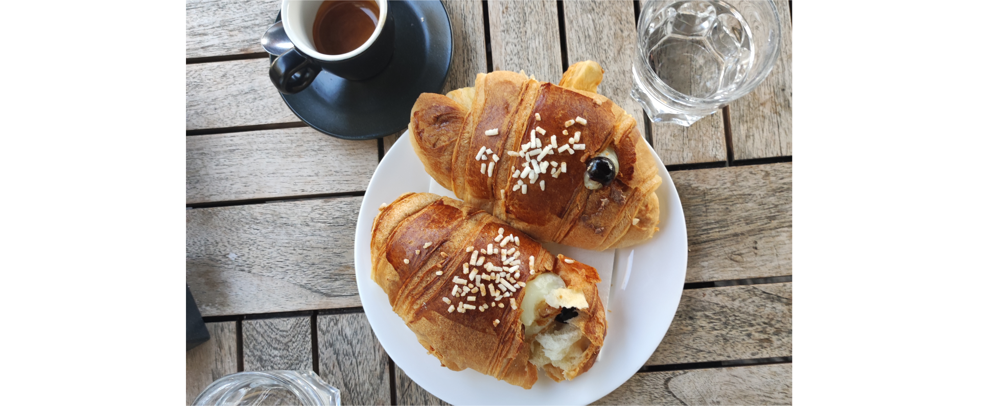 Zdjęcie przedstawia dwa słodkie wypieki przypominające nadziewane drożdżówki oraz małą filiżankę z espresso i szklankę z wodą.