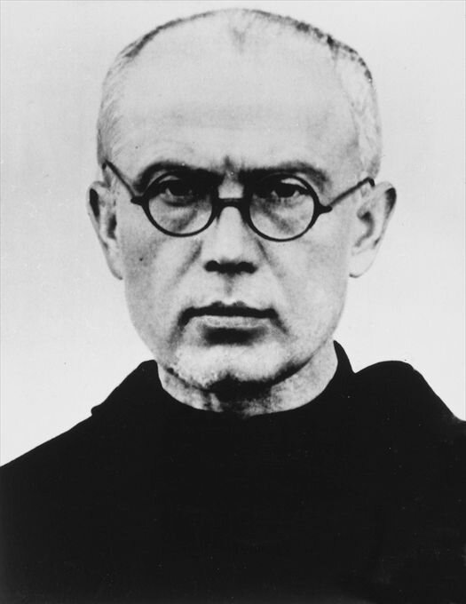Czarno‑białe zdjęcie przedstawia Maksymiliana Marię Kolbe. Ma on krótkie siwe włosy i czarne okulary. Ma na sobie czarny strój.
