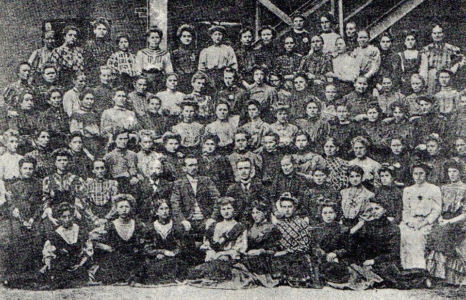 Zniszczone czarno‑białe zdjęcie przedstawia grupę kilkudziesięciu kobiet pozujących do zdjęcia. Stoją w kilku rzędach najprawdopodobniej na rusztowaniu. Są ubrane w suknie. 