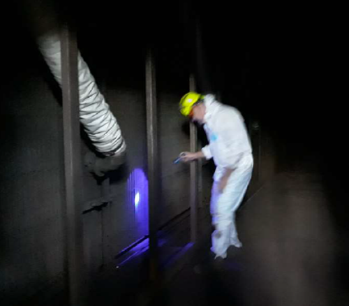 Na zdjęciu znajduje się mężczyzna ubrany w biały kombinezon oraz żółty kask, który sprawdza nieszczelności filtra za pomocą lampy UV.