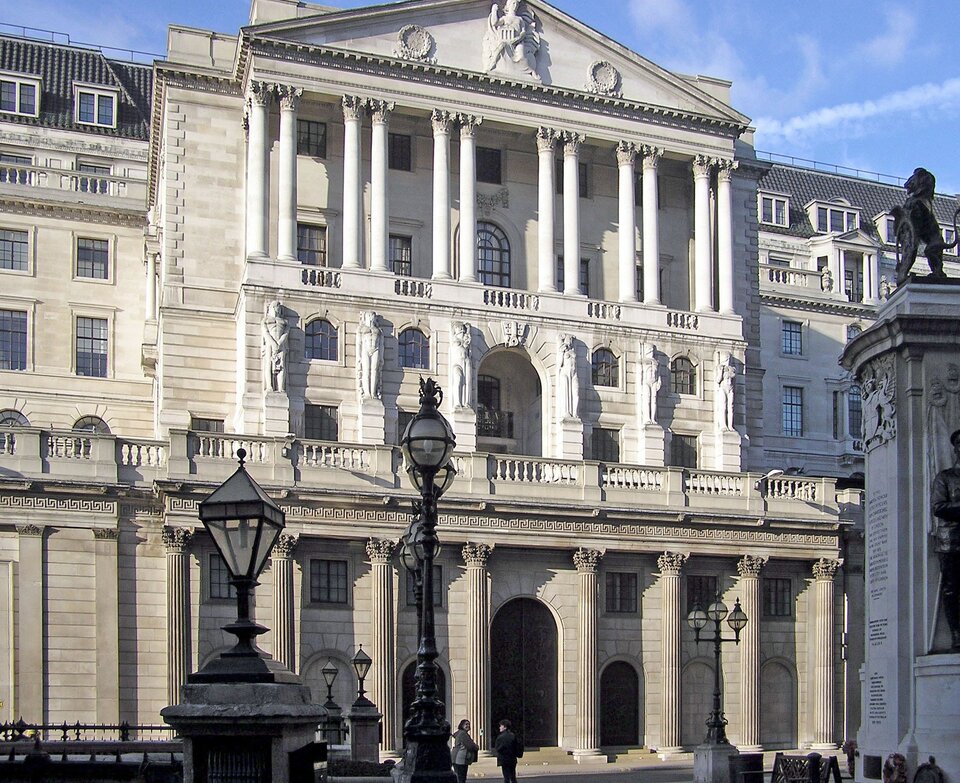 Zdjęcie przedstawia Bank of England, z angielskiego dosłownie Bank Anglii. Budynek jest biały, ma kilka pięter i zbudowany jest w stylu neoklasycystycznym. Przy wejściu stoją kilkumetrowe kolumny, nad wejściem widnieje taras, na poziomie którego znajdują się kilkumetrowej wysokości rzeźby. Na kolejnym piętrze jest dużych rozmiarów balkon, z balustradą, którą przecina sześć par kolumn. 