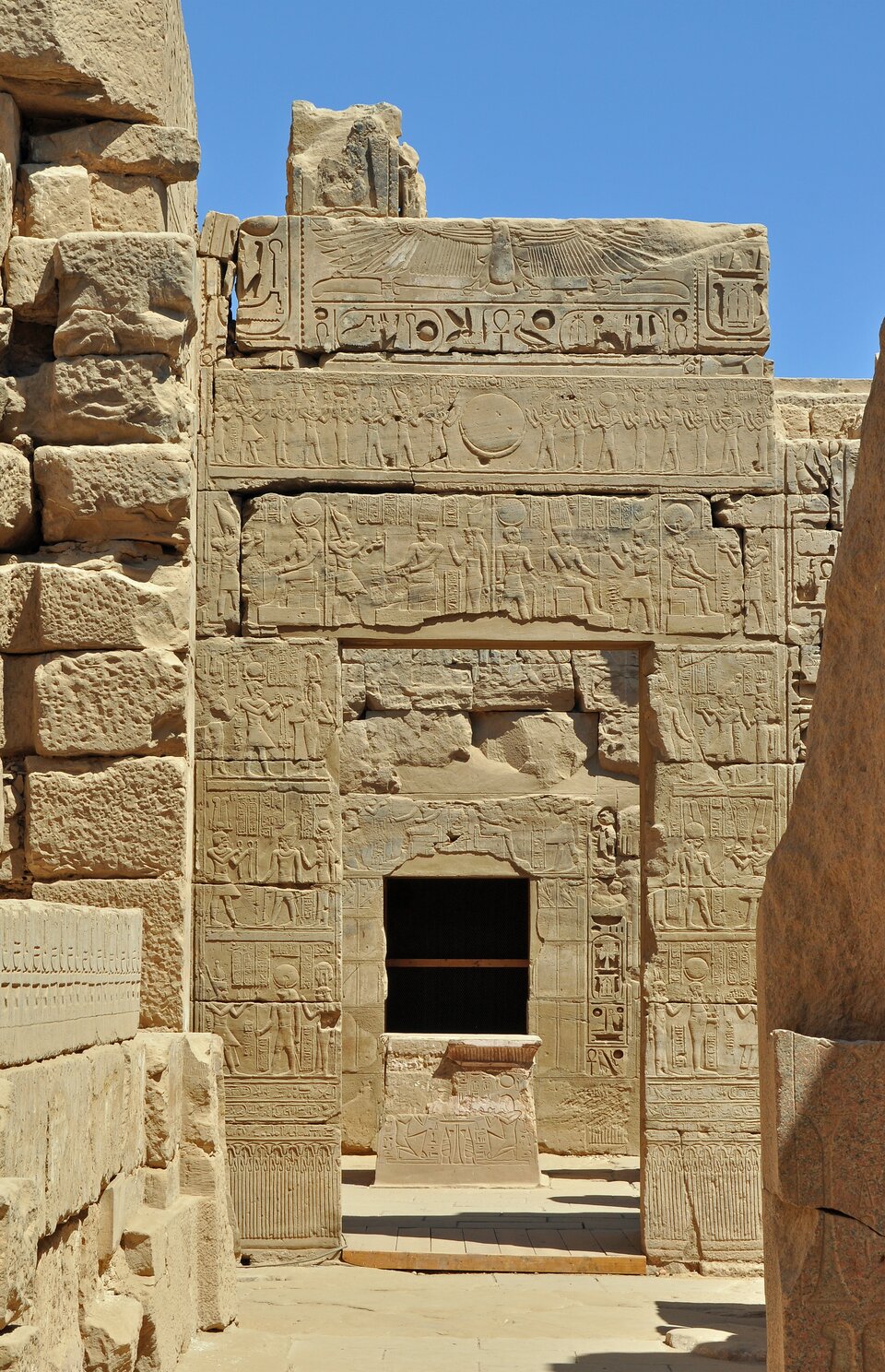 Fotografia przedstawia  jedno z wejść świątyni Chonsu w Karnaku, wykonane z jasnego piaskowca i bogato zdobione reliefami. W centralnej części kadru znajduje się monumentalny portal z prostokątnym otworem wejściowym, ponad którym rozmieszczono poziome pasy dekoracyjne. Górną partię wieńczy szeroki fryz z przedstawieniem uskrzydlonego dysku słonecznego, zwieńczonego przez symbole królewskie i hieroglify. Poniżej - kolejne pasy z reliefami figuralnymi przedstawiającymi procesje kapłanów i sceny rytualne z udziałem bóstw oraz faraona. Całość kompozycji pokryta jest hieroglifami, wypełniającymi niemal każdą wolną przestrzeń powierzchni kamiennej. U podstawy wejścia znajduje się niewielki ołtarz lub kamienny postument. Otwór wejściowy prowadzi do ciemniejszego wnętrza.