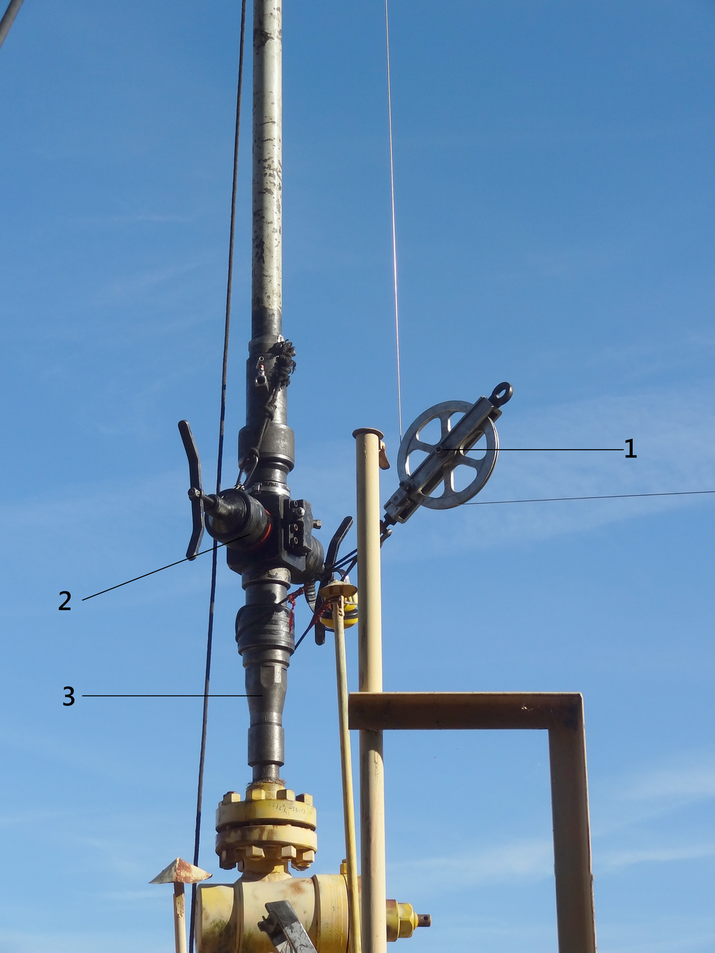 Na zdjęciu widoczny jest prewenter znajdujący się na głowicy eksploatacyjnej. Na dole zdjęcia widoczny jest fragment żółtej głowicy eksploatacyjnej. Do głowicy przy pomocy połączenia kołnierzowego przyłączony jest czarny element o kształcie pionowej podłużnej rury. Jest to prewenter. Prewenter patrząc się od dołu do góry ulega nieznacznemu rozszerzeniu. Widoczne miejsce zmiany średnicy jest oznaczone cyfrą trzy oznaczającą łącznik śluzy pomiarowej. Nad łącznikiem znajduj się zawór. Zawór jest o kształcie sześcianu. Do elementu dochodzi cyfra dwa oznaczającą prewenter pojedynczy. Z przedniej i tylnej ściany sześcianu wychodzi kurek, Kurek jest o kształcie obrotowej wajchy. Do bocznej ściany sześcianu widocznej między kurkami dochodzi stelaż z prętów na końcu którego znajduje się krążek liniowy oznaczony cyfrą jeden. Od góry sześcianu odchodzi dalsza część podłużnej rury. 