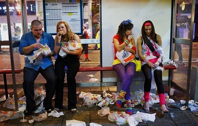 Zdjęcie przedstawia przystanek autobusowy w dużym mieście. Na ławce siedzi czworo młodych osób, jedzą fast foody. Dokoła nich leży mnóstwo śmieci.