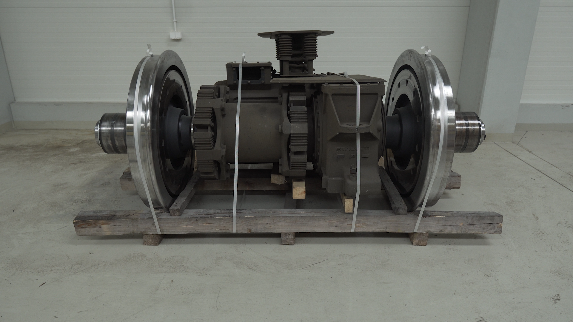 Ilustracja przedstawia silnik trakcyjny. Silnik wraz z zestawem kołowym znajduje się na palecie. Jest zabezpieczony metalowymi linkami. Silnik trakcyjny jest cylindrycznej obudowy, która została wykonana z metalu, został zamontowany między dwoma kołami kolejowymi. Silnik składa się z stojana oraz obracającego w nim wirnika.