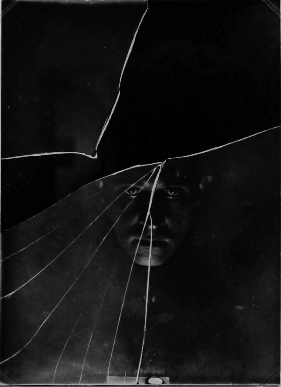 Na zdjęciu widać czarno‑białą fotografię lub portret, na którym centralnie widoczna jest ludzka twarz – oczy, nos i usta. Całość jest przysłonięta i zniekształcona przez pękniętą szybę. Linia największego pęknięcia przecina twarz niemal symetrycznie, dzieląc ją na dwie części. Twarz wydaje się niewidoczna w całości – tylko fragmenty są oświetlone.