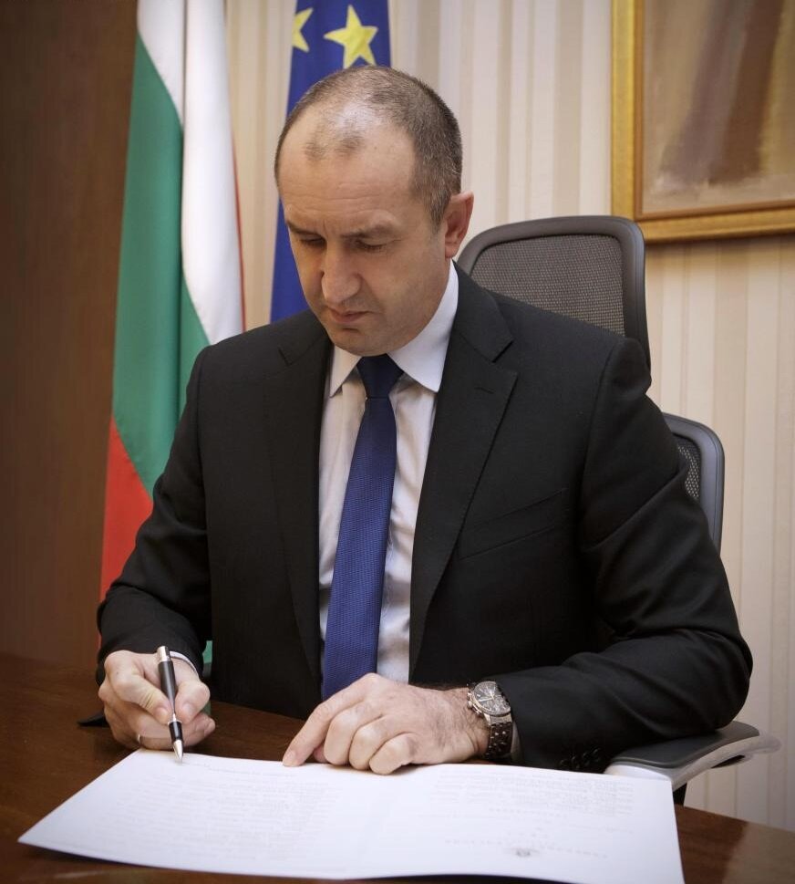 Zdjęcie przedstawia dojrzałego mężczyznę w garniturze. Mężczyzna siedzi przy biurku i podpisuje dokumenty. Za jego plecami widać dwie flagi: Bułgarii i Unii Europejskiej.
