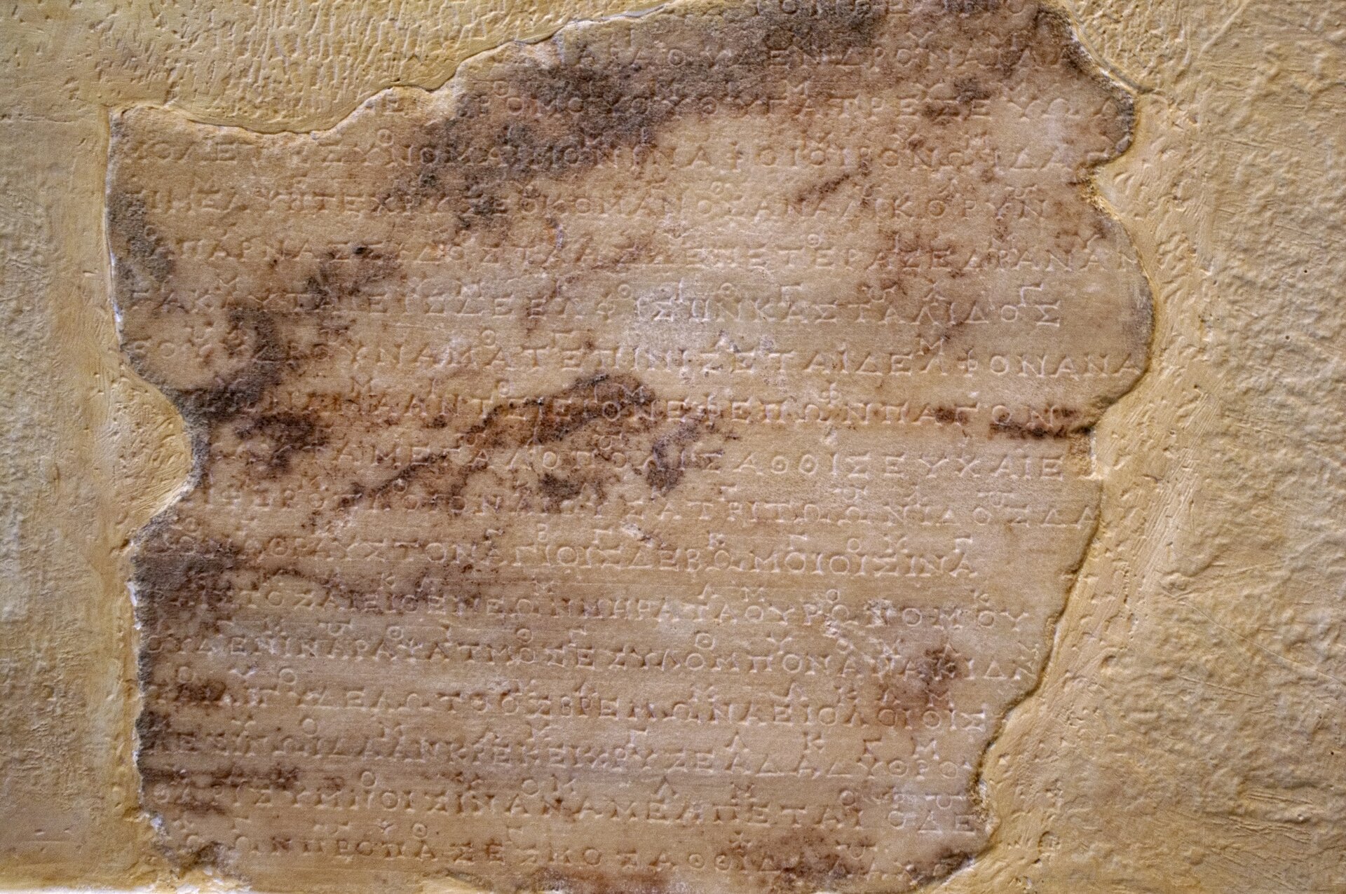 Ilustracja przedstawia tekst w języku greckim wyryty w ścianie z jasnego kamienia. Kamień z wyrytym napisem, w  kolorze beżowym z brązowymi wzorami, jest otoczony ścianą w jaśniejszym, kolorze. 