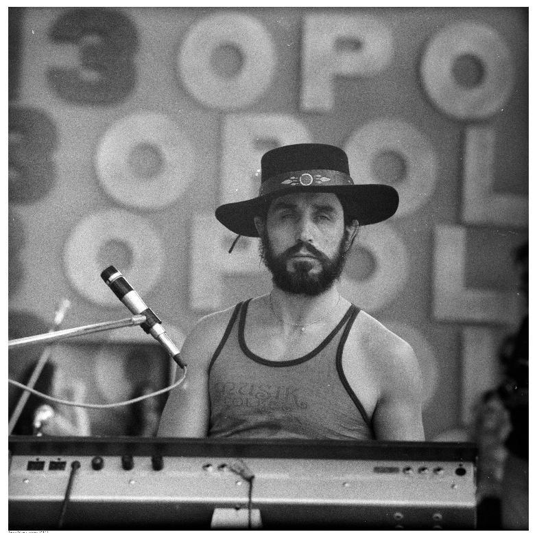 Czarno‑białe zdjęcie przedstawia mężczyznę grającego na keyboardzie. Ma brodę, nosi ciemny kapelusz z ozdobnym paskiem i koszulkę bez rękawów z napisem. Na pierwszym planie widoczny jest mikrofon, a w tle duże litery układające się w słowo „OPOLE”, sugerujące festiwal muzyczny. Scena ma charakter koncertowy.