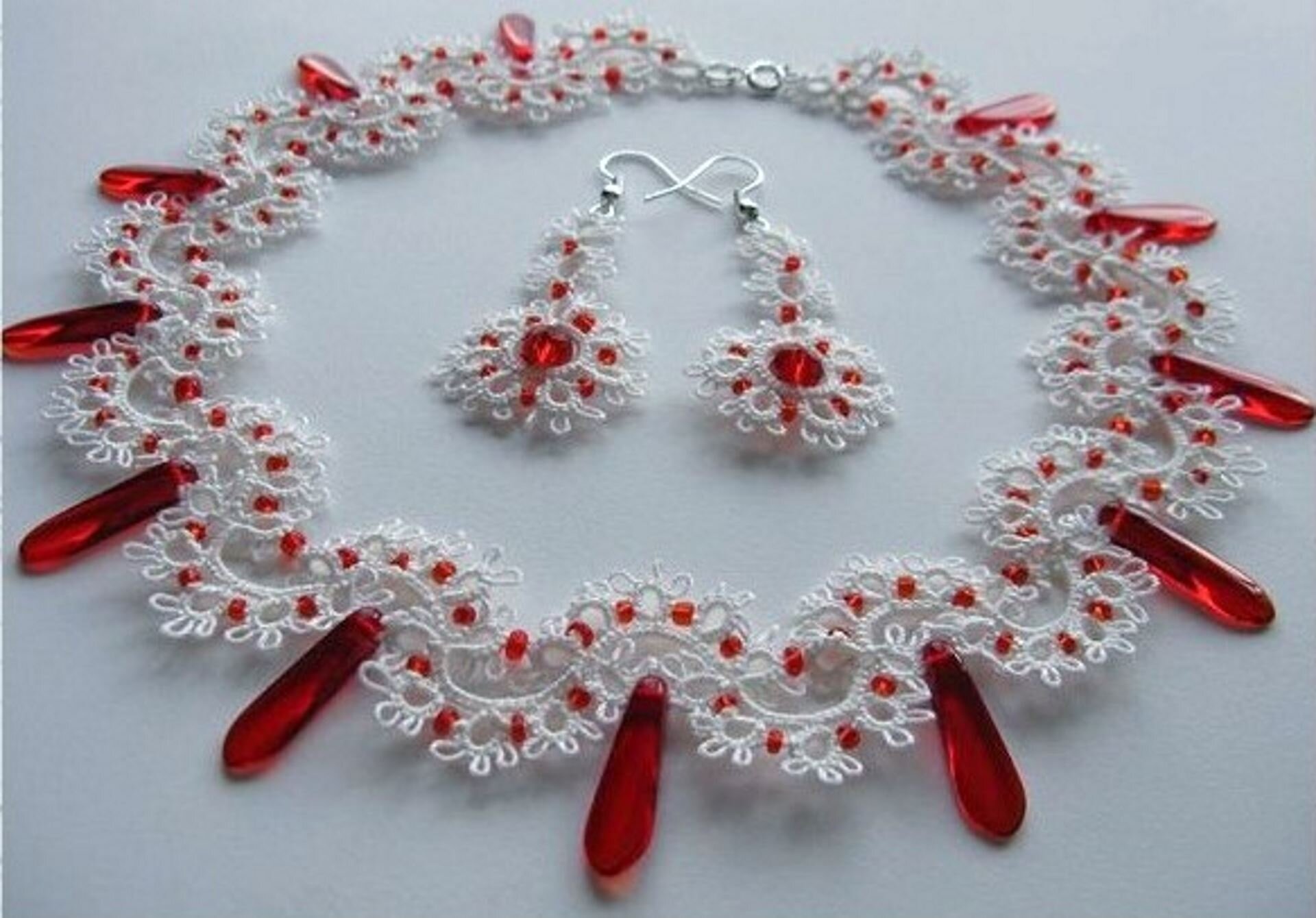 Ilustracja przedstawia zdjęcie ręcznie wykonanej biżuterii frywolitkowej (naszyjnik i kolczyki), rodzaj koronki artystycznej. Naszyjnik i kolczyki leżą na białej tkaninie. Do jej wykonania wykorzystano białą koronkę, którą połączono z czerwonymi, przeźroczystymi kamieniami ozdobnymi. Naszyjnik składa się z koronkowych kwiatków układających się w falowany okrąg. Między kwiatkami znajdują się drobne, czerwone koraliki. Do, co drugiej, fali kwiatków przyczepiono podłużne, czerwone, szklane korale. Na środku leżą dwa kolczyki w kształcie kwiatków, w środku każdego znajduje się okrągły, czerwony koralik, płatki kwiatków również ozdobione są mniejszymi, czerwonymi koralikami.