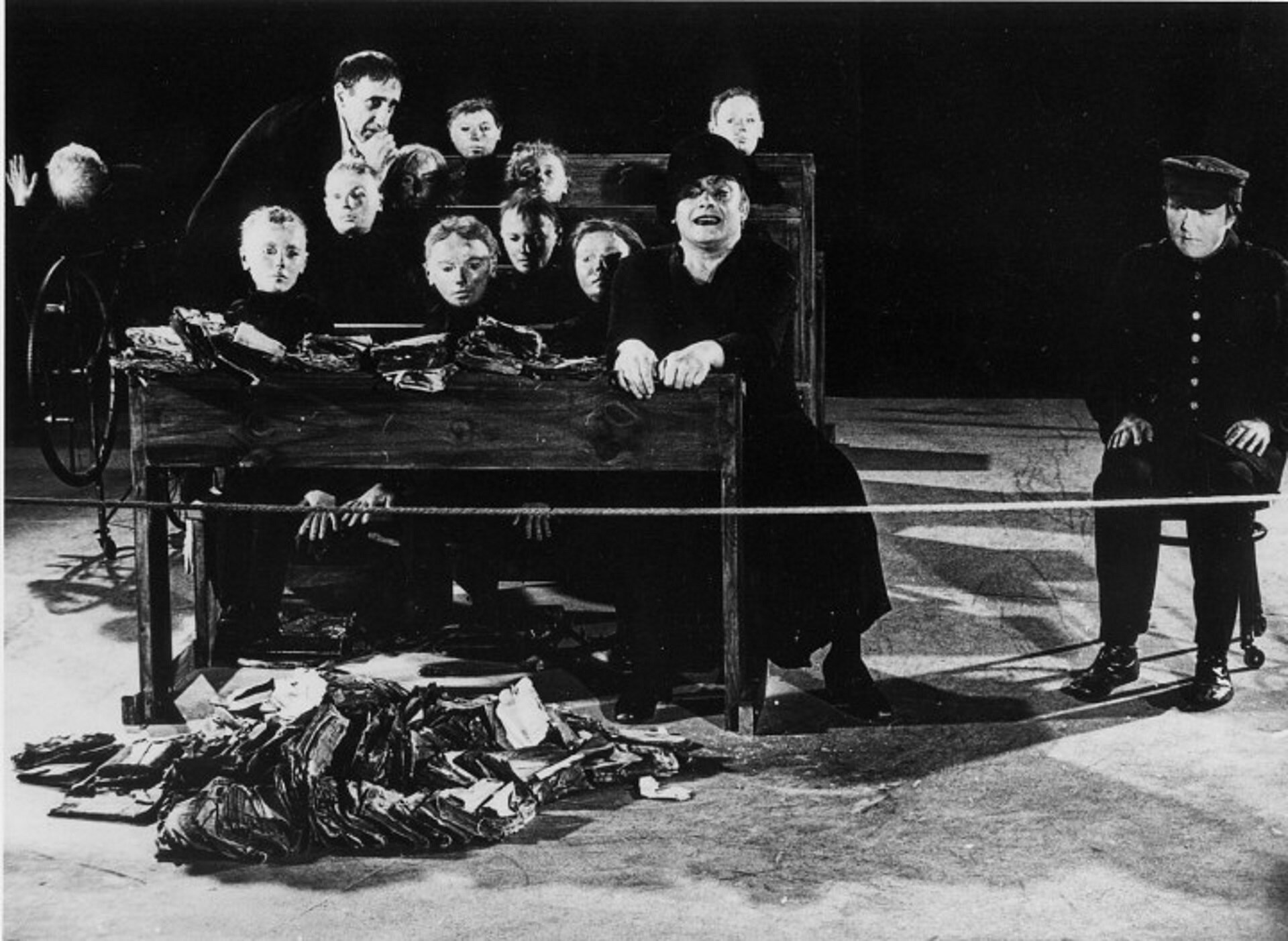 Ilustracja przedstawia czarno-białą fotografię, która przedstawia aktorów na scenie, ubrani są wszyscy na czarno, mają smutne miny. Aktorzy odgrywali wolę w spektaklu „Umarła klasa”.