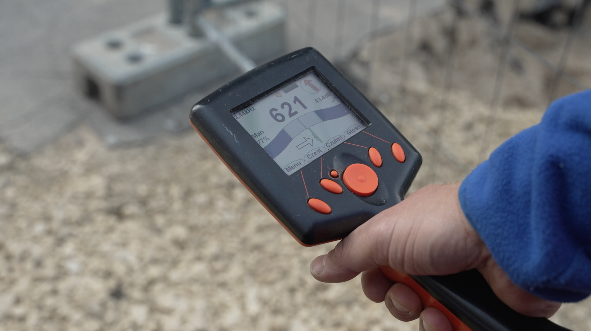 Na grafice pracownik trzymający w dłoni detektor do wykrywania infrastruktury podziemnej w trakcie pracy. Na jego ekranie wyświetlają się wyniki pomiarów. W tle kamyki i siatka.