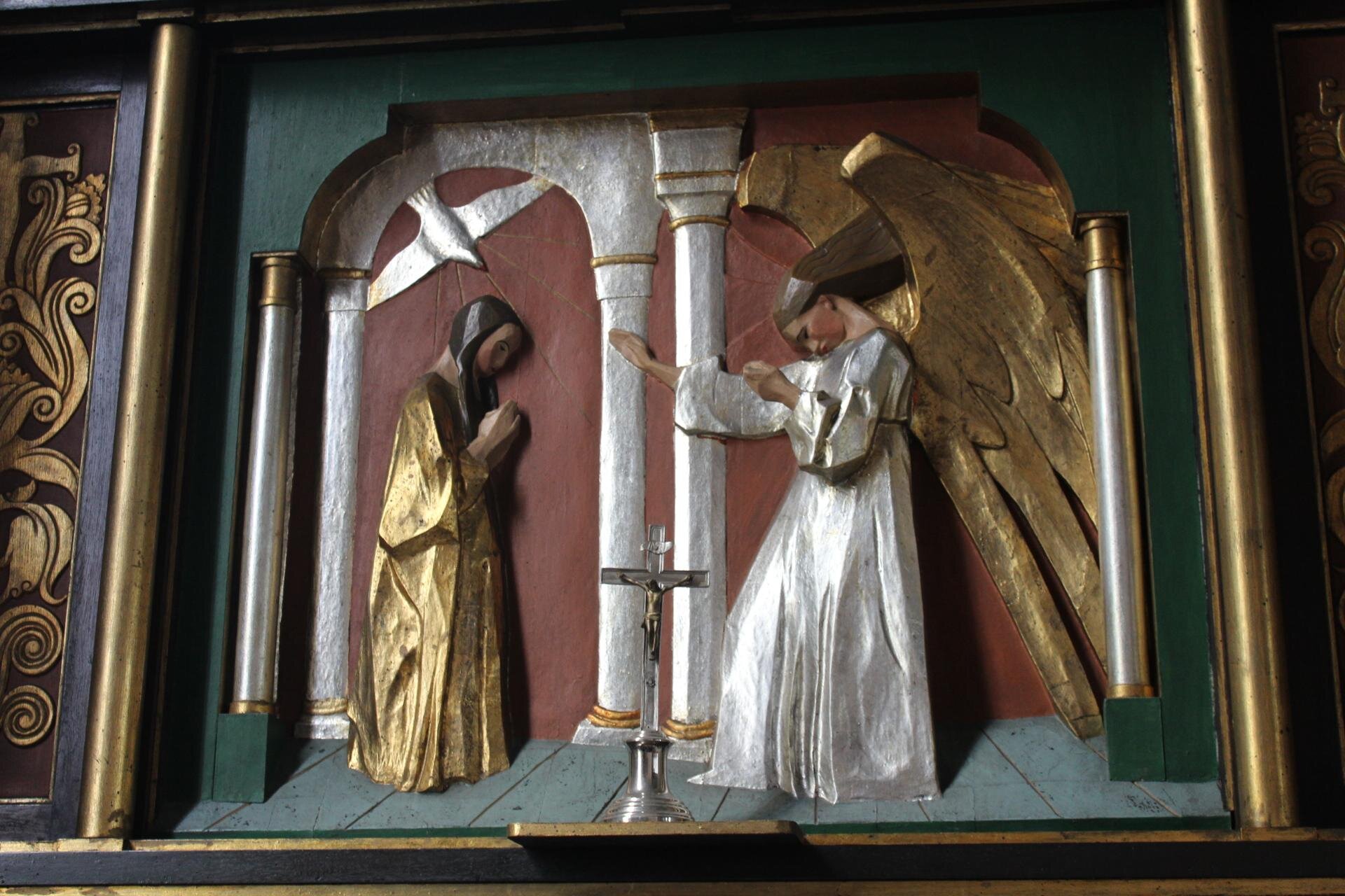 Zdjęcie przedstawia płaskorzeźbę ukazującą anioła i kobietę. Z lewej strony, w złotej sukni i długich, brązowych włosach, stoi bokiem kobieta. Ma złożone dłonie i pochyla głowę. Nad nią znajduje się biały ptak. Przed nią stoi, bokiem do widza, anioł w białej sukni, długich, brązowych włosach i dużych, złotych skrzydłach. Pomiędzy postaciami dwie kolumny oraz krzyż stojący na ziemi. Płaskorzeźba oprawiona jest w ciemnozieloną ramę, wykończoną złotem. 