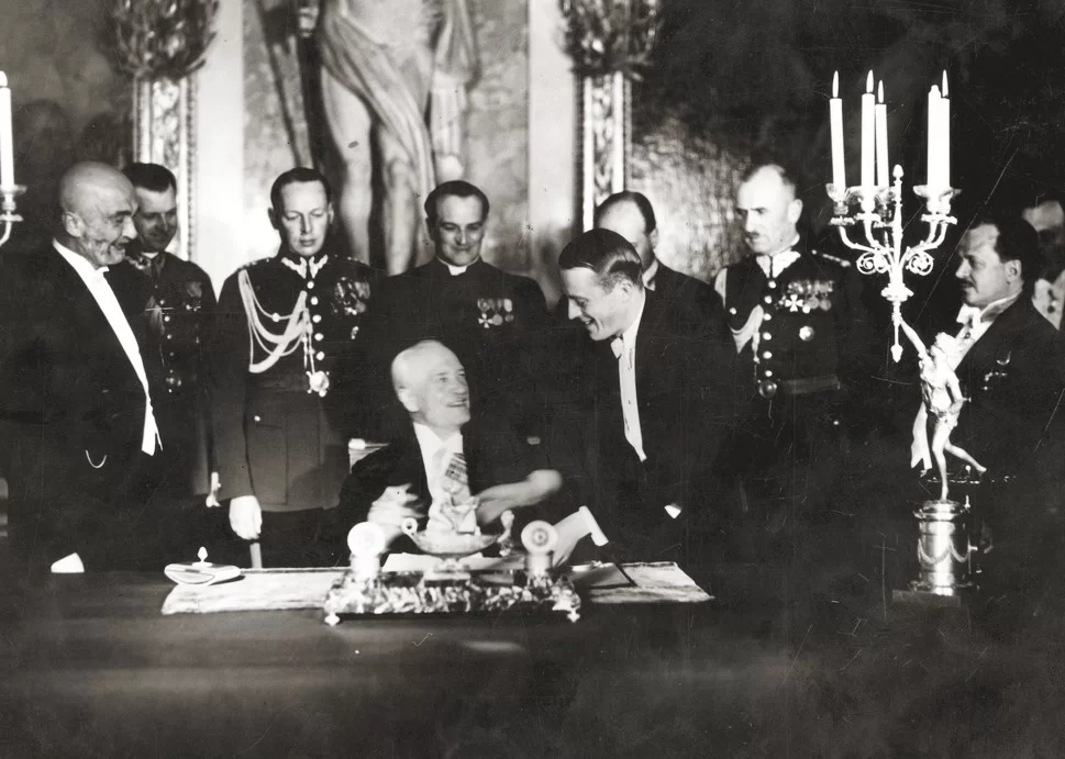 Fotografia przedstawia grupę mężczyzn. Jeden z nich, Ignacy Mościcki siedzi przy stole, przed nim leży dokument. Prezydent jest uśmiechnięty. Wokół niego stoją odświętnie ubrani pozostali mężczyźni. Ubrani są w mundury, na których znajdują się liczne odznaczenia wojskowe. Jeden z nich podaje Mościckiemu pióro. Podpisanie Konstytucji ma miejsce w sali, której ściany zdobione są licznymi posągami oraz obrazami.