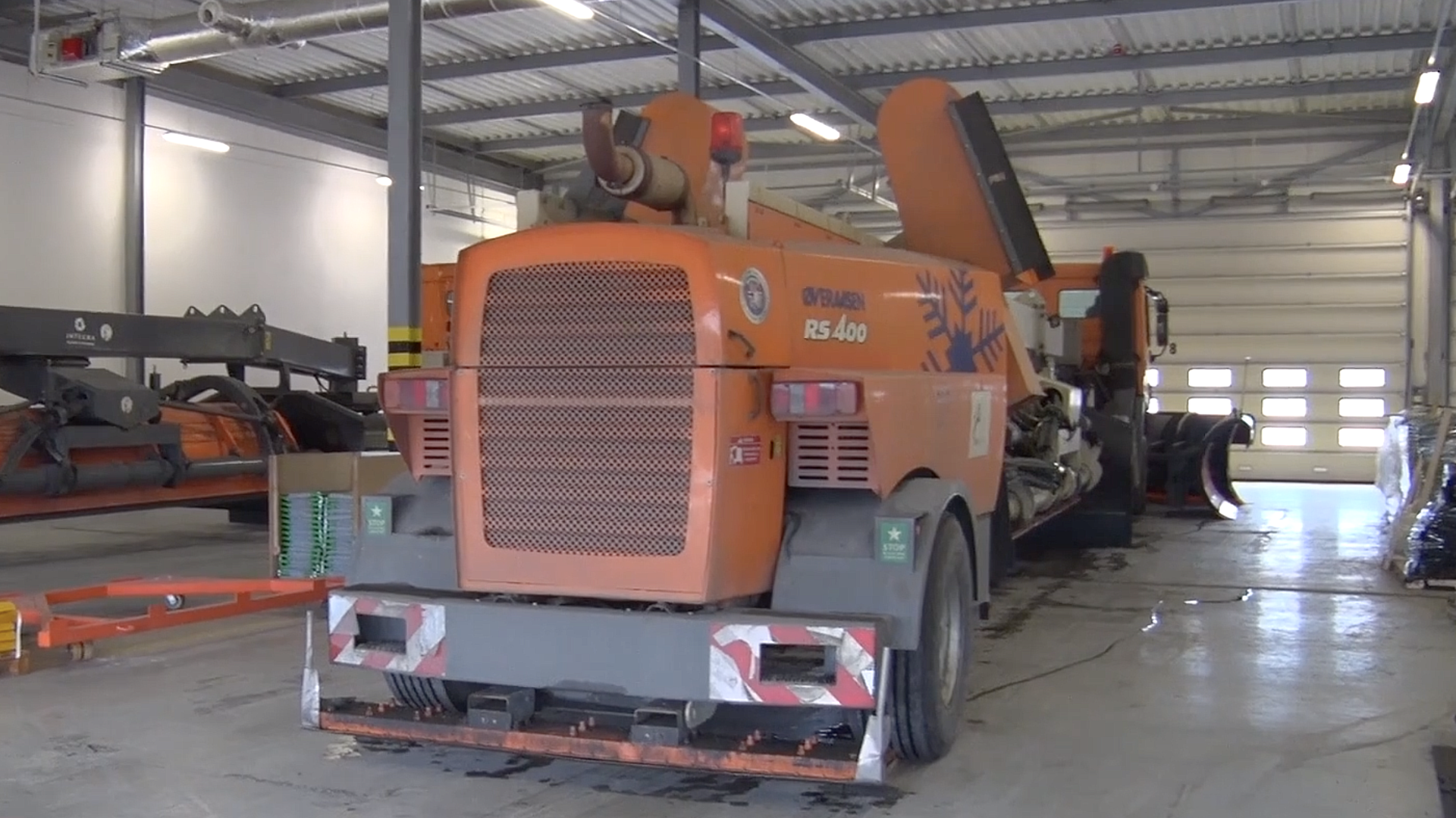 Zdjęcie przedstawia polewarko‑zmywarkę. Jest to duży ciężki pojazd przypominający traktor. Z tyłu maszyny znajduje się długa metalowa płaska konstrukcja. Polewarko‑zmywarka ma zamontowaną z przodu na górze odpowiednio wygiętą metalową rurę zwróconą przed pojazd. Służy ona do wypuszczanie odpowiednich mieszanek chemicznych po to, aby oczyścić lub odśnieżyć chemicznie powierzchnię płyty lotniskowej.