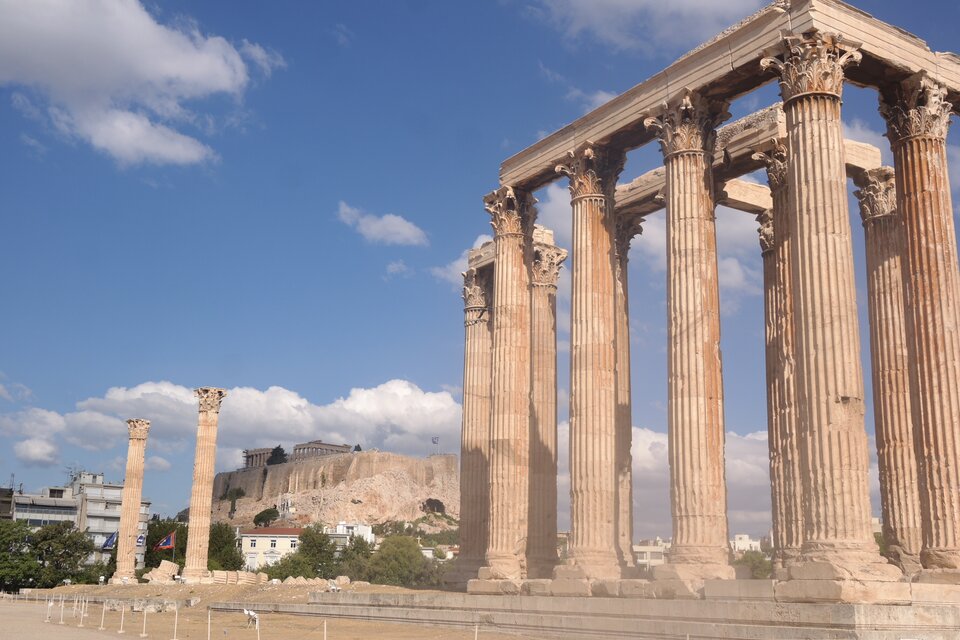 Zdjęcie przedstawia ruiny świątyni Zeusa Olimpijskiego w Atenach. W prawej części zdjęcia widocznych jest kilka kolumn korynckich. Są one zwieńczone głowicami z bogato rzeźbionymi liśćmi akantu. W lewej części zdjęcia, w oddali, widać jeszcze dwie takie kolumny. Stoją one samotnie. Tło ruin stanowi ukazany w oddali Akropol, a także współcześnie wybudowane budynki mieszkalne.