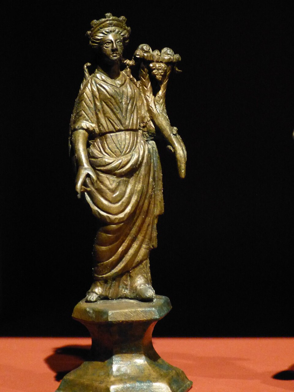 Zdjęcie przedstawia posąg młodej kobiety w długiej, udrapowanej sukni. Kobieta trzyma przy lewym ramieniu uniesioną dłonią róg obfitości.