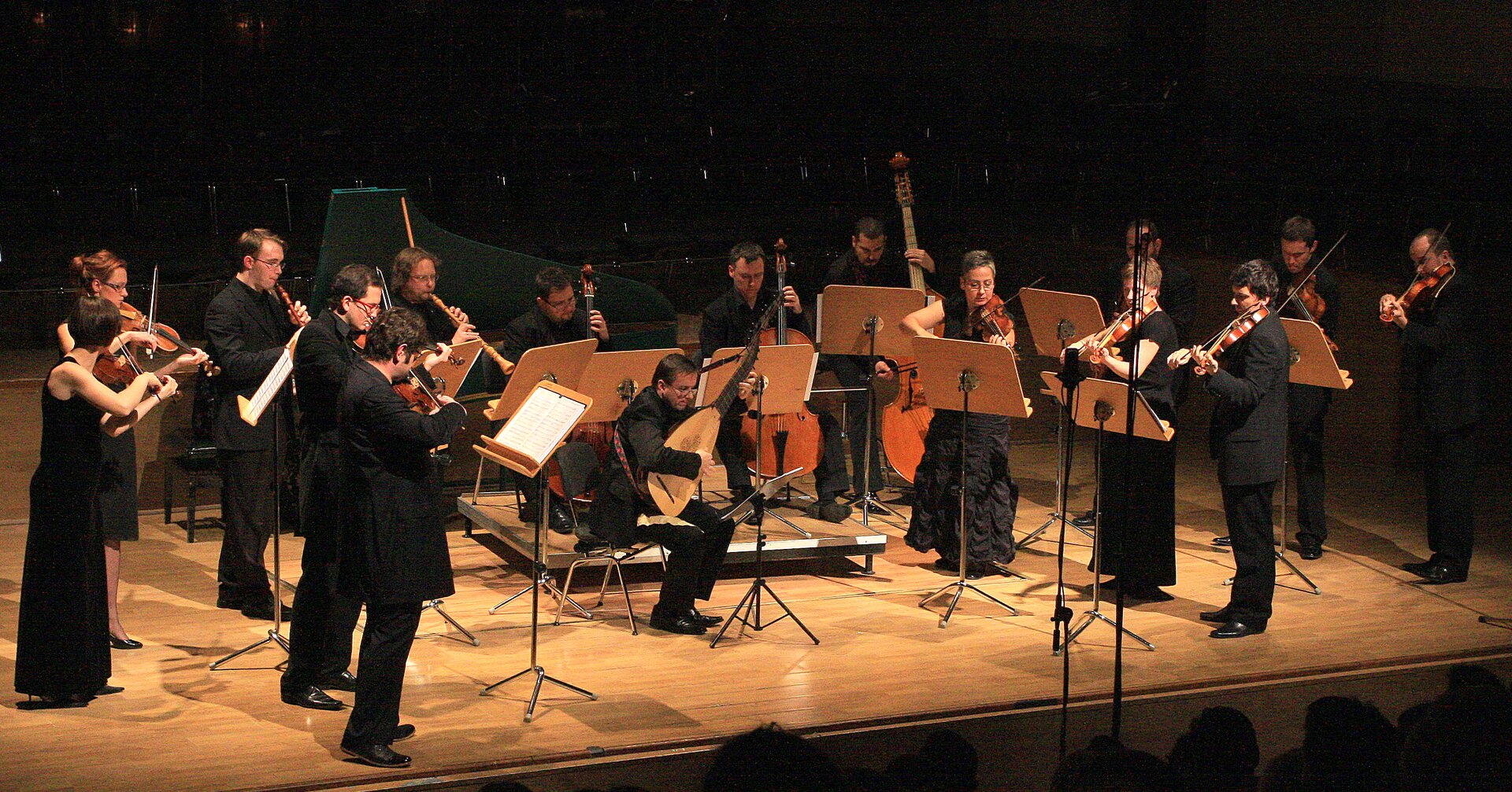 Fotografia z koncertu. Muzycy są ubrani na czarno i znajdują się na drewnianej scenie. Przed sceną rysują się zaciemnione sylwetki widowni. Muzycy ustawieni są w kształt półkola. Po bokach mniejsze instrumenty smyczkowe, z tyłu większe. Na środku siedzi mężczyzna grający na instrumencie strunowym, podobnym do gitary. Z tyłu stoi fortepian. Tło jest ciemne, oświetlona jest tylko scena.