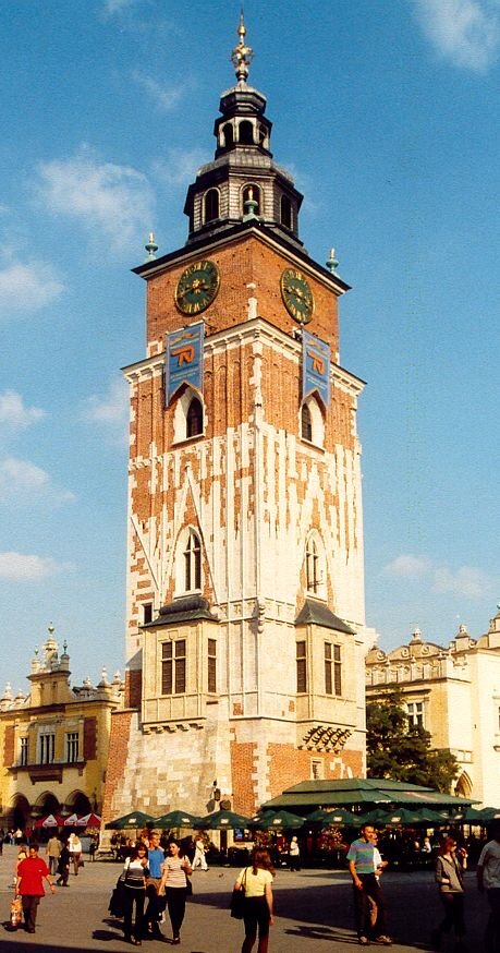 Fotografia przedstawia wysoką kwadratową wieżę, stojącą na jednym z placów. Jest zbudowana z cegieł o dwóch różnych kolorach. W górnej części na każdej ze ścian widać zegary, patrząc wyżej jest coraz węższa i zakończona ostrym czubkiem. Na placu przed wieżą spacerują ludzie. Widać też stoliki i krzesła zadaszone parasolami.