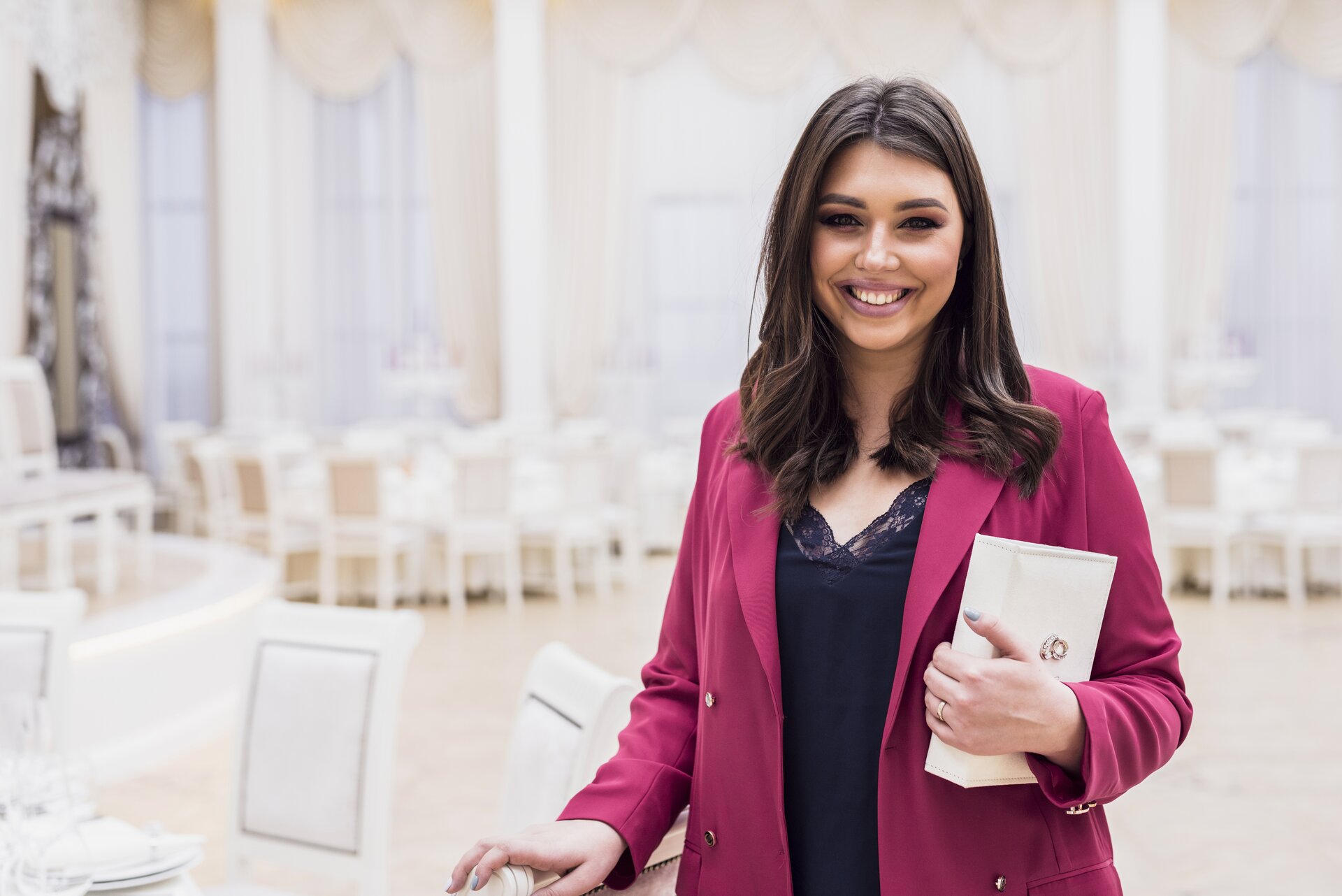 Zdjęcie przedstawia uśmiechniętą młodą kobietę. W tle widoczna sala bankietowa.