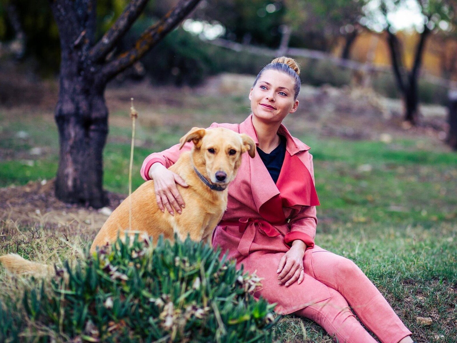 Zdjęcie przedstawia młodą kobietę z psem siedzących na trawie w parku. Kobieta ubrana jest w różowy komplet składający się z płaszcza i spodni, włosy ma upięte w koczek. Prawą dłonią obejmuje średniej wielkości psa o sierści w kolorze jasno brązowym. 