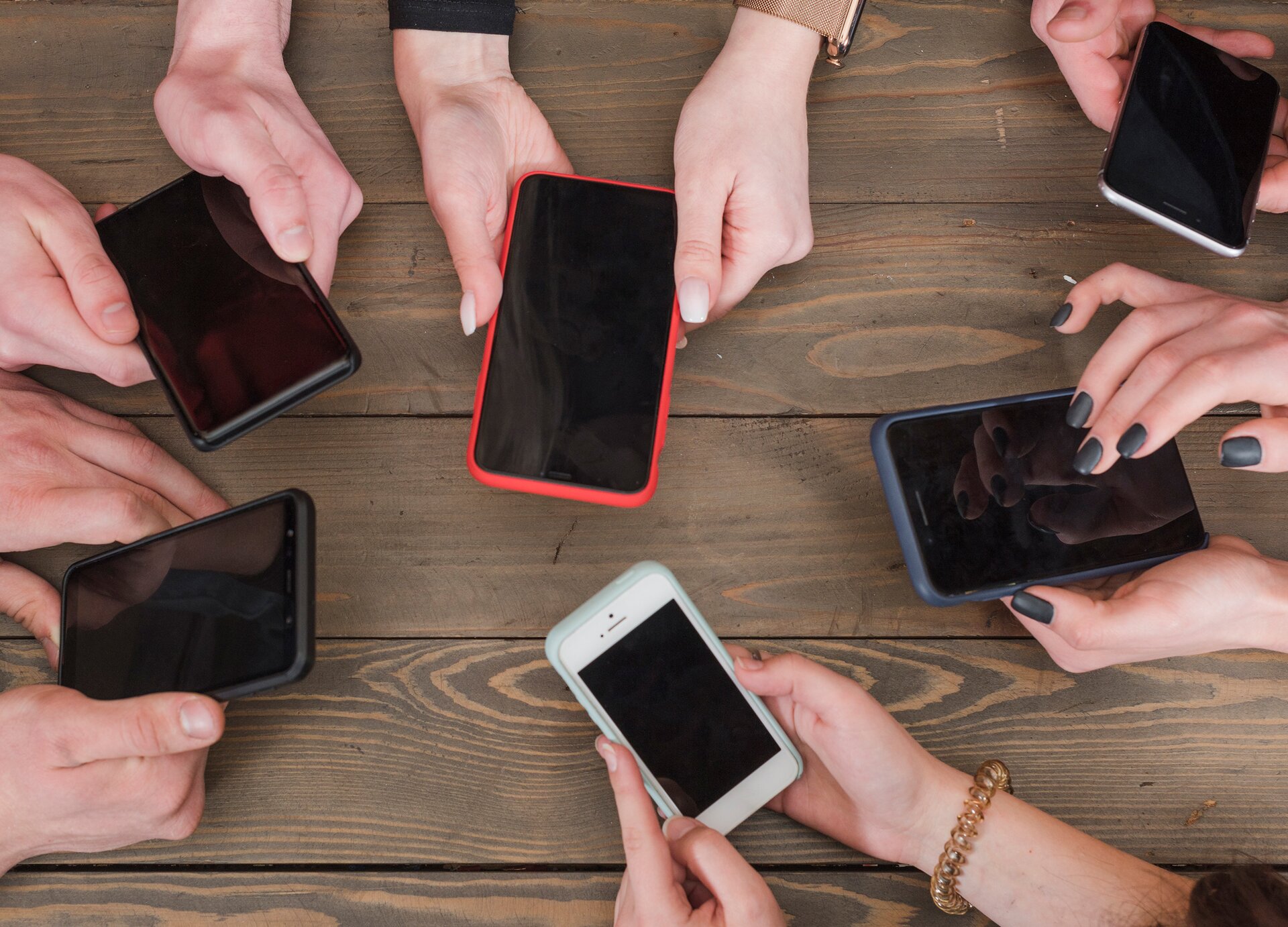 Zdjęcie przedstawia sześć par dłoni damskich i męskich, ułożonych w kole i opierających się na drewnianym blacie. Każda para rąk trzyma telefon komórkowy. Widoczne są różne modele i kolory telefonów.
