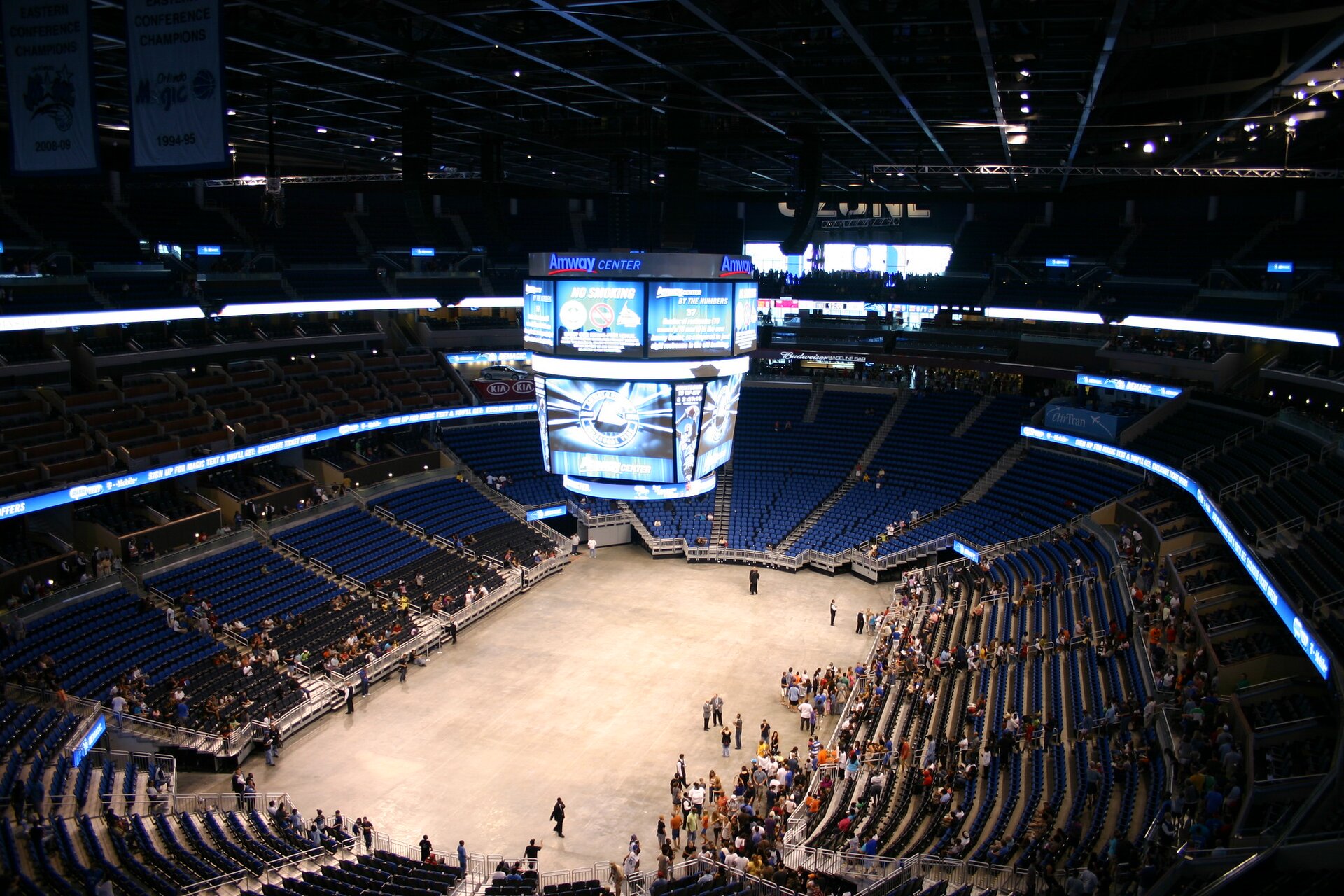 Zdjęcie przedstawia widok hali sportowej. Na trybunach znajdują się nieliczne grupy widzów. Nad podłogą hali zawieszony jest zestaw ekranów.