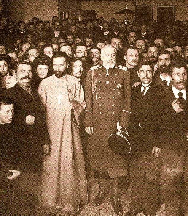 Zdjęcie przedstawia tłum ludzi. Na pierwszym planie stoi mężczyzna w długiej szacie. Na szyi ma powieszony krzyż. Mężczyzna ma czarne włosy i czarną brodę. Jest młody. Obok niego stoi mężczyzna w średnim wieku w wojskowym mundurze. Ma siwe włosy i siwą brodę. W ręce trzyma wojskową czapkę. Tłum wokół nich to głównie mężczyźni.    