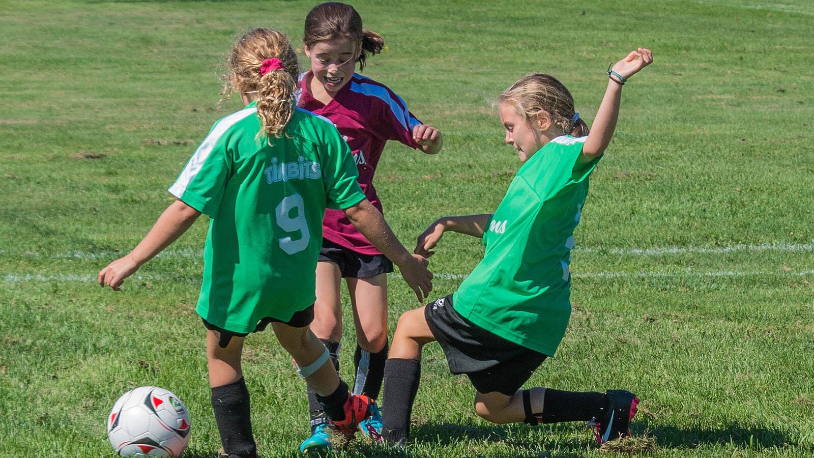 Zdjęcie przedstawia trzy dziewczynki, w wieku około 7 lat, grające w piłkę nożną na murawie. Dziewczynki ubrane są w piłkarskie stroje. Dwie z nich są ubrane w zielone koszulki i czarne szorty, a trzecia w bordową koszulkę i czarne szorty. Dziewczynki walczą o piłkę leżącą obok nich. 