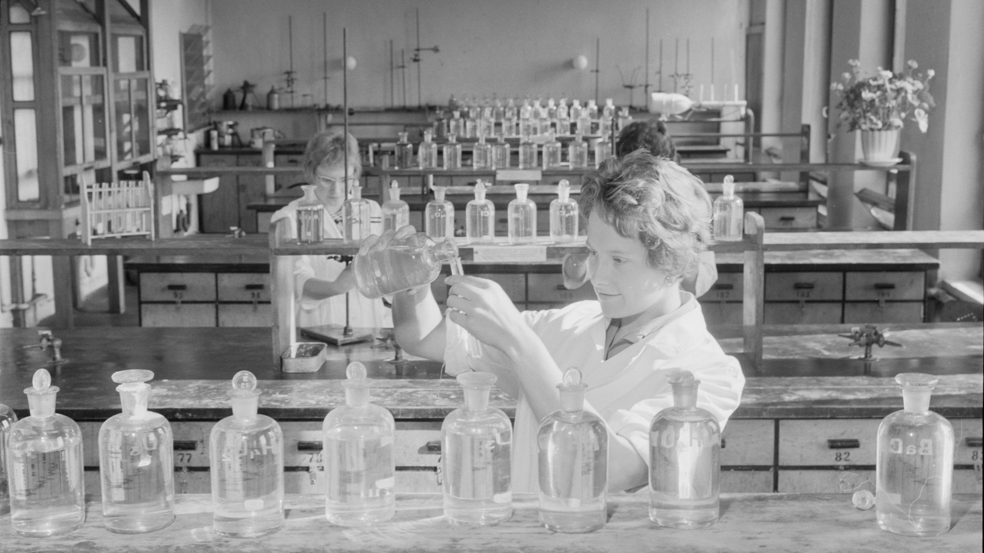 Zdjęcie przedstawia laboratorium chemiczne. Jest to duże o przestronne pomieszczenie. Znajdują się półki, oszklone szafki i stoły, na których stoją szklane butelki z odczynnikami chemicznymi. Nad stołami zamontowane jest silne oświetlenie. Widoczne są trzy kobiety wykonujące eksperymenty chemiczne.