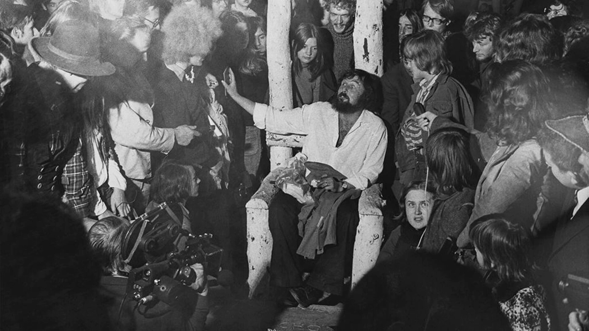 Na środku fotografii znajduje się mężczyzna, który siedzi na drewnianym krześle. Ma on ciemne włosy do ramion, wąsy i brodę. Ubrany jest w jasną koszulę z podwiniętymi do łokcia rękawami, ciemne, szerokie spodnie i ciemne buty. Na lewym nadgarstku ma zegarek, a na palcu serdecznym lewej dłoni obrączkę.  Na kolanach trzyma kurtkę. Jego lewa dłoń leży na udach, a prawa jest wyciągnięta w bok. W dłoni trzyma płaski przedmiot. Dookoła mężczyzny znajduje się tłum ludzi. Postać w lewym, dolnym rogu fotografii trzyma kamerę. 