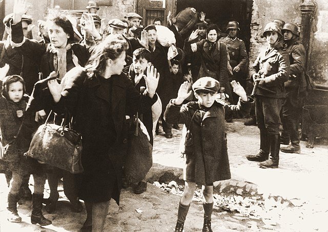 Fotografia czarno-biała, archiwalna, przedstawiająca polskich Żydów złapanych przez Niemców podczas tłumienia powstania w warszawskim getcie. Scena dzieje się na ulicy, przed budynkiem. Grupa Żydów, głównie kobiet i dzieci, stoi odwrócona plecami do żołnierzy niemieckich, którzy są w gotowości do strzału. Żydzi podnoszą ręce do góry na znak poddania. 