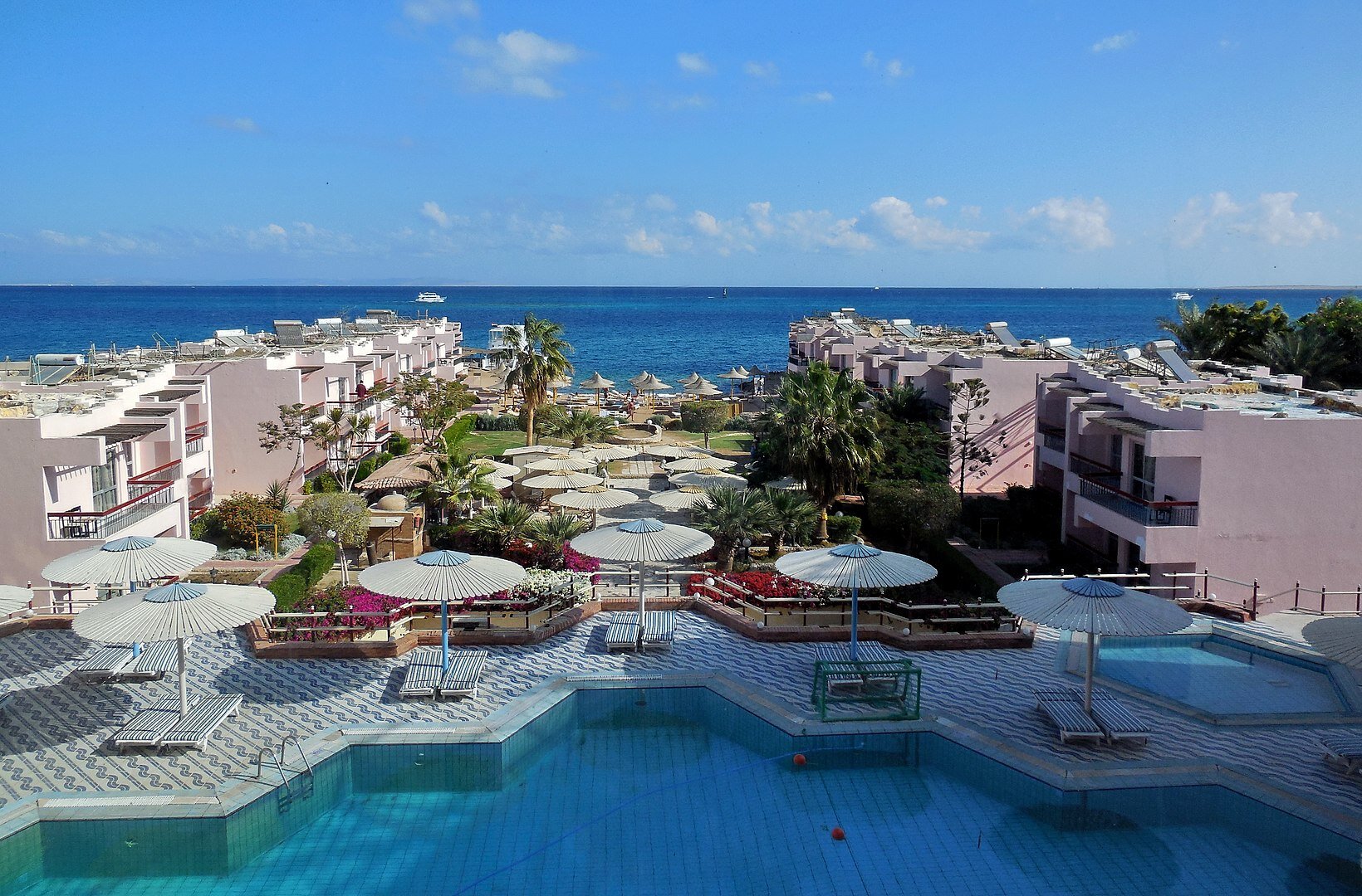 Zdjęcie przedstawiające przestrzeń nadmorskiego kurortu. Na pierwszym planie rozległy basen, obok leżaki i parasole. Dalej teren zielony, a po bokach niskie zabudowania hotelowe. W tle morze.