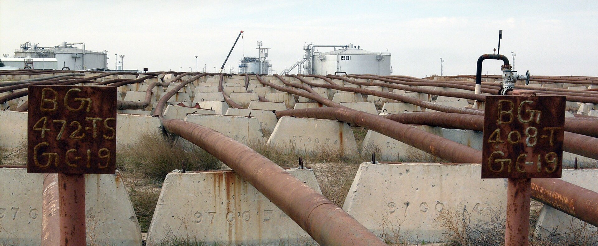 Zdjęcie przedstawiające rurociągi o średnicy kilkudziesięciu centymetrów położone na betonowych wspornikach około pół metra nad ziemią. Rurociągi kierują się w stronę dużych, okrągłych zbiorników na drugim planie.